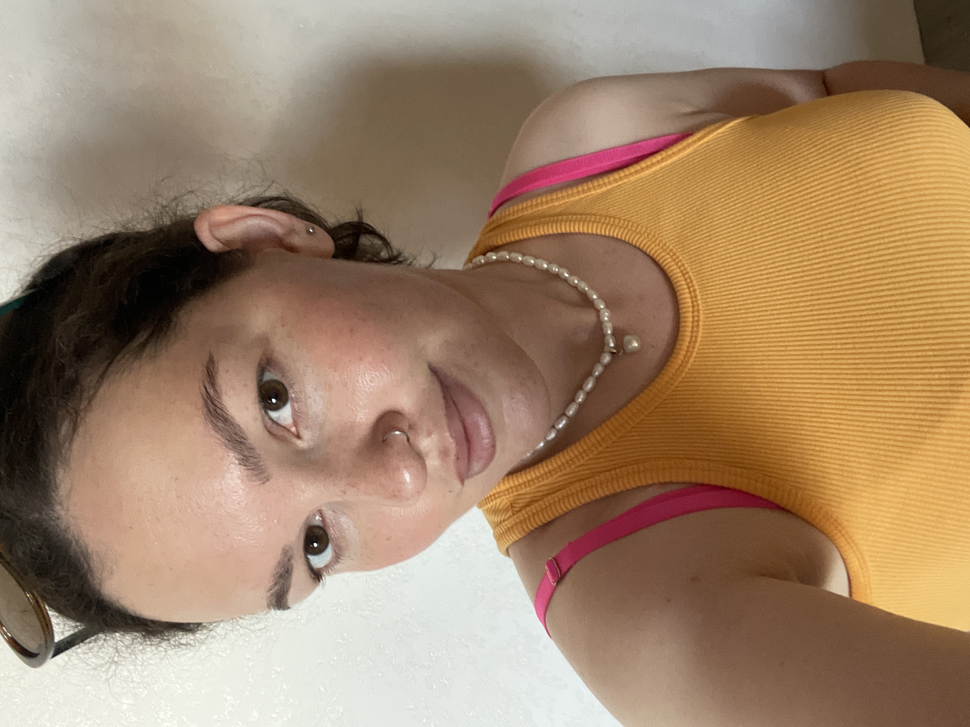 A woman is pictured in a close-up shot. She has fair skin, dark brown hair, and brown eyes. She is wearing an orange ribbed tank top with thin pink straps and a pearl necklace. There is a nose ring in her left nostril and an earring in her left ear. The background appears to be a plain white wall. The image provides no information about the location being in the city of Izhevsk, Russia.