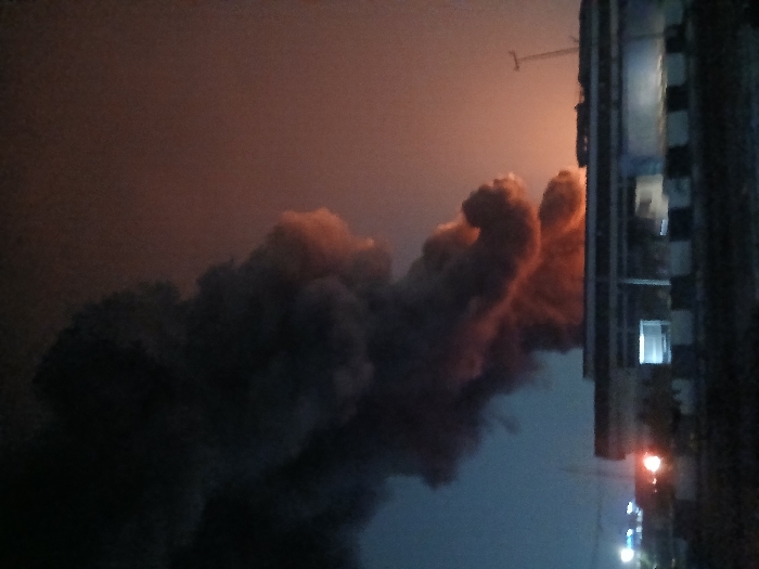 Thick plumes of dark smoke fill the frame, tinged orange by flames. A tall building stands to the right, with lit windows punctuating its facade. Streetlights glow faintly below, suggesting a nighttime setting. The overall scene hints at a fire or major incident, casting a dramatic and unsettling atmosphere. The sky behind the smoke has an eerie orange glow.