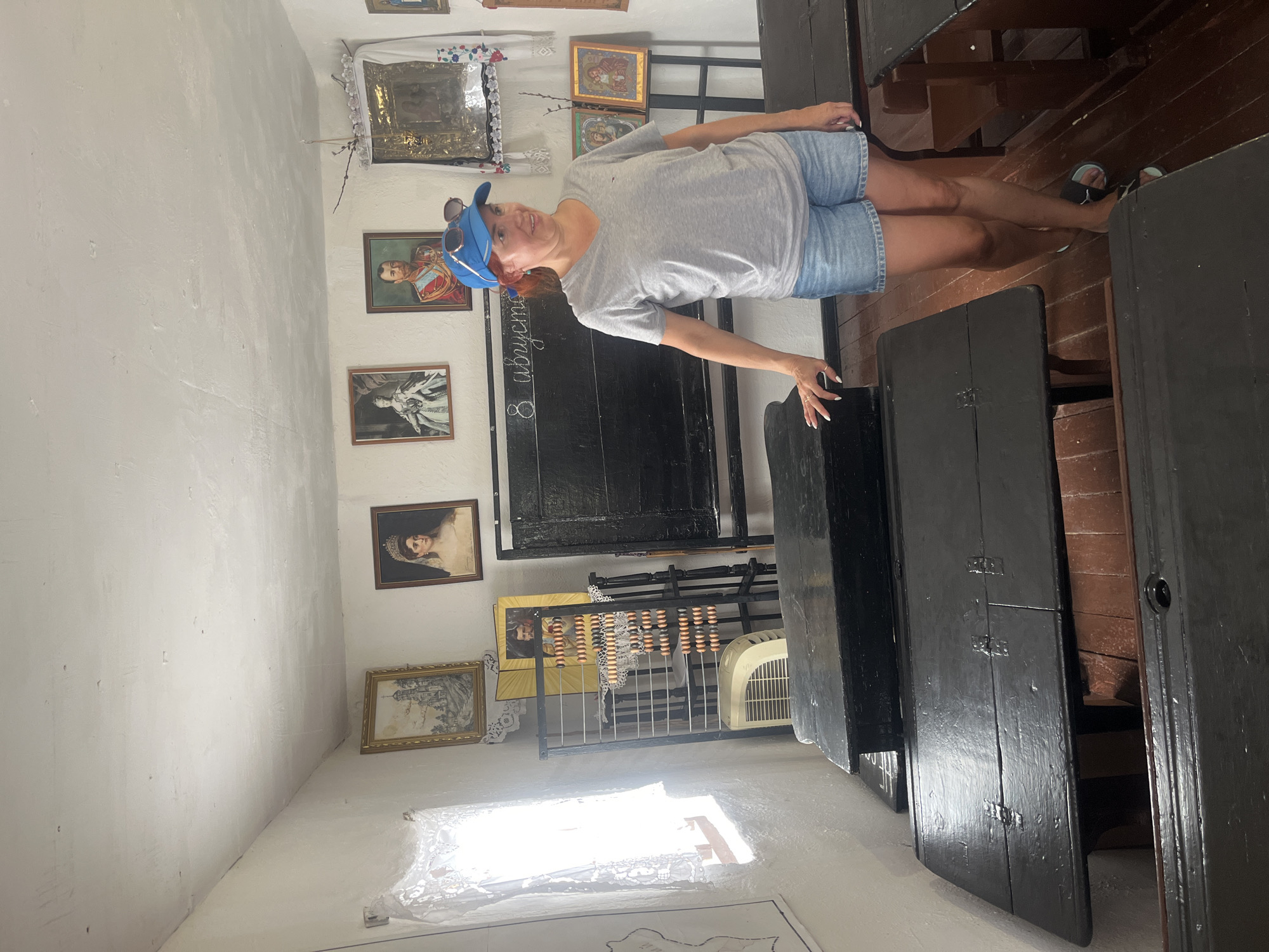 A woman stands in a rustic classroom in Taman, Russia. She wears a grey t-shirt, denim shorts, and a blue cap, with sunglasses perched on the cap. The classroom features antique wooden desks and a chalkboard with writing on it. Portraits of historical figures hang on the walls, along with religious icons. A window with lace curtains lets in natural light. An abacus and an old fan are also visible. The overall atmosphere is one of historical preservation and education.