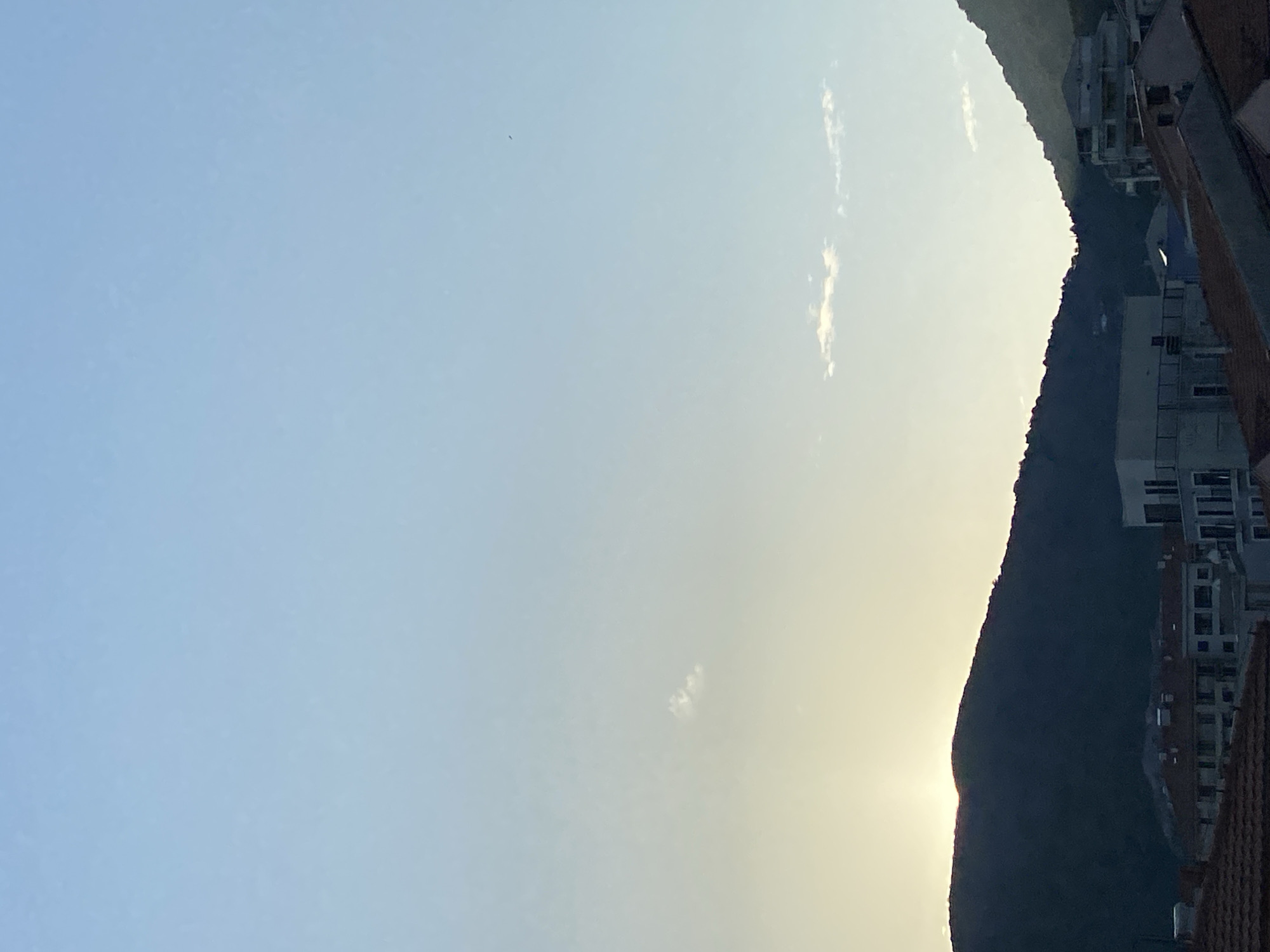 The image shows a wide shot of a mountainous landscape behind a city, likely taken from a slightly elevated position looking towards the coast. The sky is a gradient of pale blue at the top, transitioning to a soft yellow and then white near the horizon, suggesting either sunrise or sunset. A faint sliver of the sun is visible peeking over the dark silhouette of a mountain range in the lower left quadrant.
Several buildings with red tiled roofs and modern white structures are visible in the mid-ground, forming part of the city of Budva. The buildings are densely packed, indicating an urban environment. In the background, the dark forms of mountains dominate the scene, their peaks softened by the atmospheric conditions. A few sparse clouds are scattered across the sky, with one small, wispy cloud just above the center of the frame and a slightly larger, more defined cloud formation in the lower right section of the sky. There are no people or animals visible in the frame. The overall impression is one of a serene, early morning or late evening view of a coastal city nestled against a mountainous backdrop.