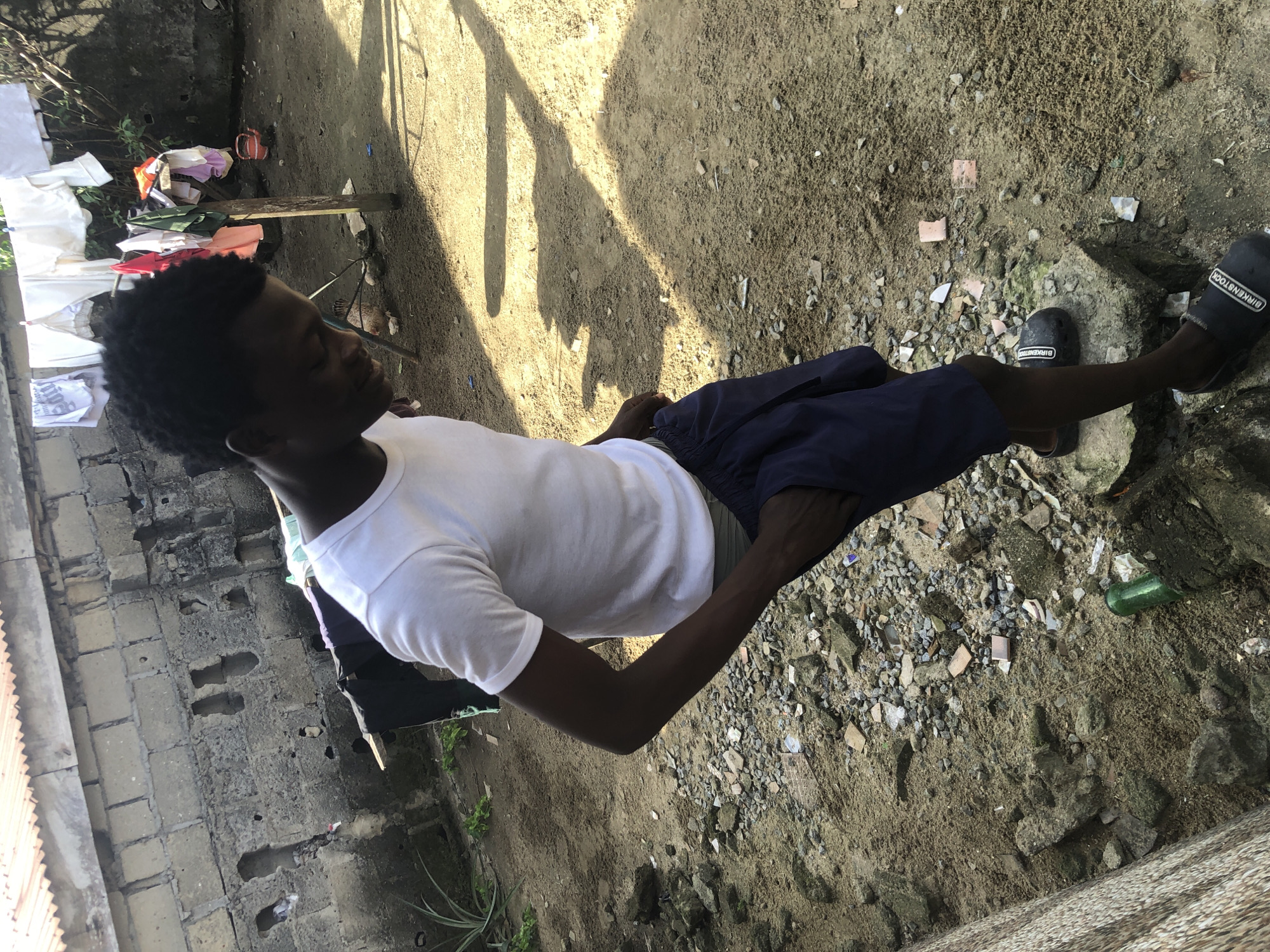 The image depicts a young Black male wearing a white t-shirt and dark blue shorts, standing outdoors in what appears to be an urban or semi-urban environment. He is looking down and has one hand in his pocket. The ground is covered in sand, dirt, and scattered rocks and debris, with some small pieces of broken tile. In the background, there is a partially constructed brick wall with some areas of damage and missing sections. Clothes are hanging on a line strung between wooden poles, with a variety of garments visible, including white shirts, underwear, and a red kettle on the ground. A small chicken is visible near the clothesline. The lighting suggests it is daytime, with strong shadows cast on the ground. The scene has a gritty, weathered aesthetic, potentially indicative of a developing area or a location in need of upkeep. The presence of a green glass bottle on the ground and a rough-looking wall suggest a possibly impoverished or rural setting. The location is identified as Ajah, Nigeria.