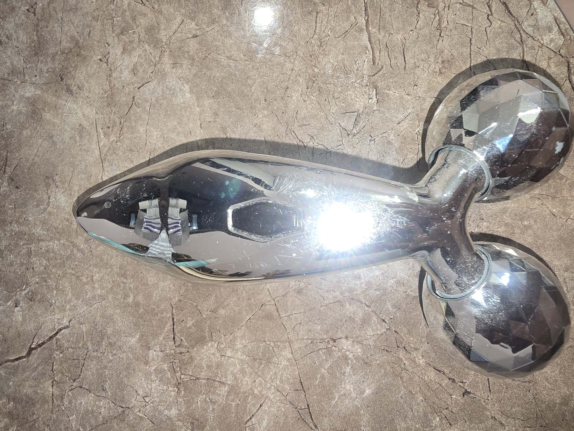 A silver-colored facial roller, shaped like a small dumbbell with two faceted, spherical ends and a hexagonal indentation in the middle of its handle, rests on a textured, marble-like surface. The surface has a brown and beige mottled pattern with dark veins resembling cracks. The roller exhibits scratches and smudges, and is heavily illuminated by a bright light source, creating strong reflections and glare, particularly on the lower spherical ends and the central hexagonal area. The lighting suggests the photograph was taken indoors, with artificial light being the primary source. The scene is static and does not depict any activity or people. The location in Ulan-Ude, Russia, is not visually discernible from the provided image.