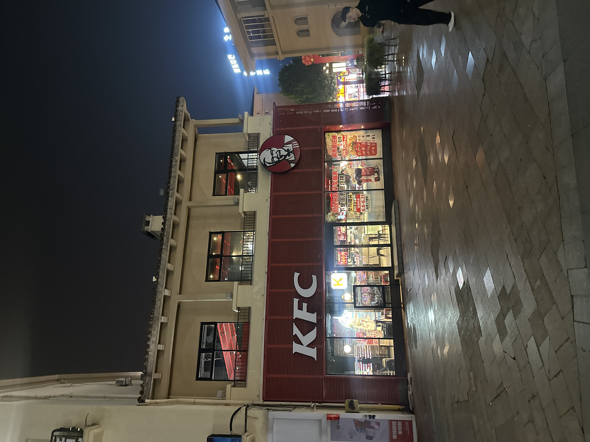 The image depicts a KFC restaurant located in the city of Chongqing High-tech Zone, China. The establishment is a two-story building with a red facade featuring the KFC logo and Colonel Sanders' portrait. The ground floor is a storefront with large glass windows displaying the interior and promotional materials. The pavement in front of the restaurant is wet, suggesting recent rain, and is paved with rectangular stones. A person in dark clothing is walking on the pavement to the right of the frame. Other buildings and streetlights are visible in the background, indicating an urban setting at night.