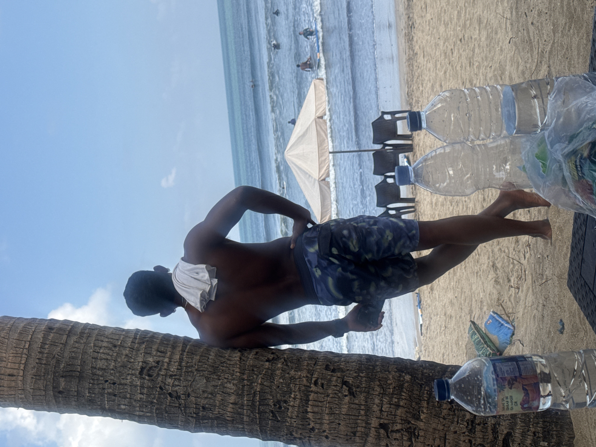 A man stands leaning against a palm tree on a sandy beach in Kuta, Indonesia. He is wearing camouflage swim trunks and has a white towel draped over his shoulders. The man is facing away from the camera, looking out at the ocean where several people are surfing. In the foreground, two plastic water bottles and a bag of snacks sit on a dark surface. Beach chairs and a large umbrella are visible further down the beach. The sky is blue with scattered clouds.