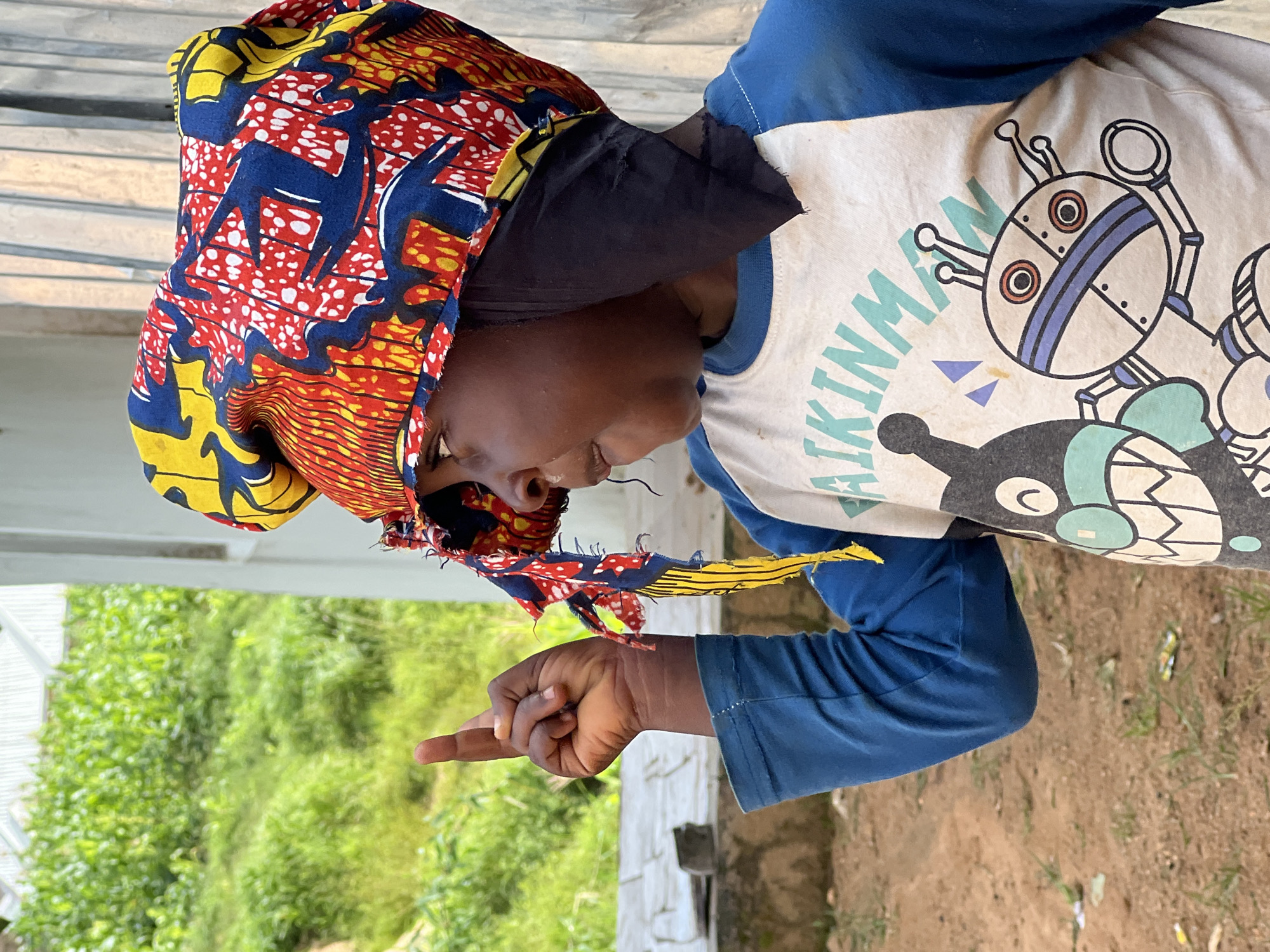 In Jos, Nigeria, a young child wears a vibrant, colorful headwrap of red, yellow, and blue batik fabric. They are also wearing a blue long-sleeved shirt with a graphic print on the front featuring a cartoon robot and text. The child is raising their right hand, index finger pointed upwards. They are looking off to the right, with their mouth slightly open. The background consists of a building with vertical siding and lush green foliage, suggesting an outdoor setting in a rural or semi-urban area. The ground is composed of dry earth and scattered debris. The image is a medium shot, focusing on the child and their attire, with the environment serving as a backdrop.