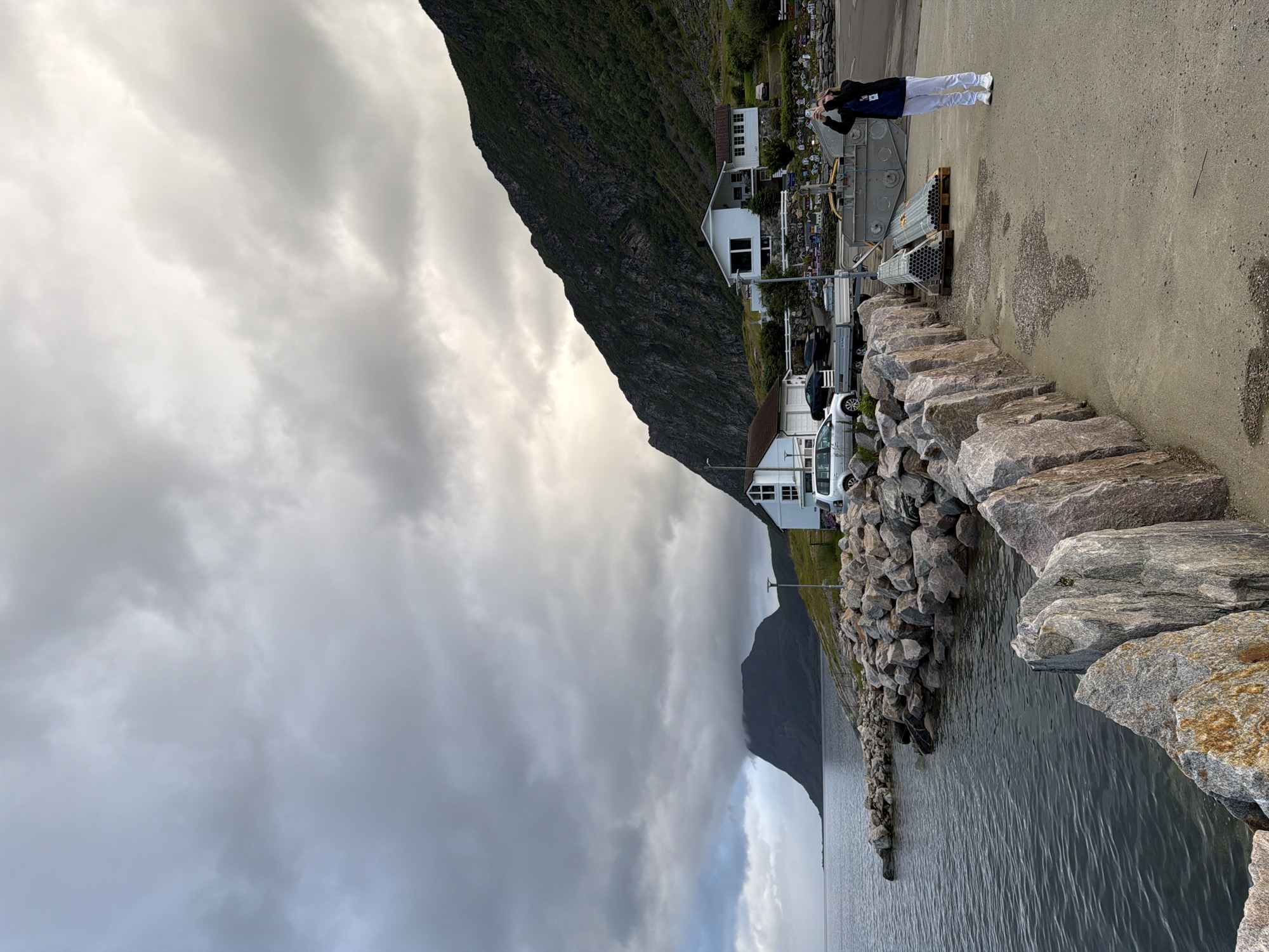The image depicts a cloudy day scene in Harstad, Norway, by the water. In the foreground, large gray and reddish-brown rocks line the edge of a concrete pier. To the right, a person stands, wearing a dark jacket, blue shirt, and white pants, holding up a phone to take a picture. Behind them, stacked metal pipes on wooden pallets are visible, along with a large gray industrial container. Further back, a white house with dark windows is situated on a grassy slope leading up to a steep, dark green mountain. Several cars are parked near the house and on a road. In the distance, the calm, dark water of the sea extends to the horizon, where a large, dark mountain peak, partially obscured by clouds, rises majestically.