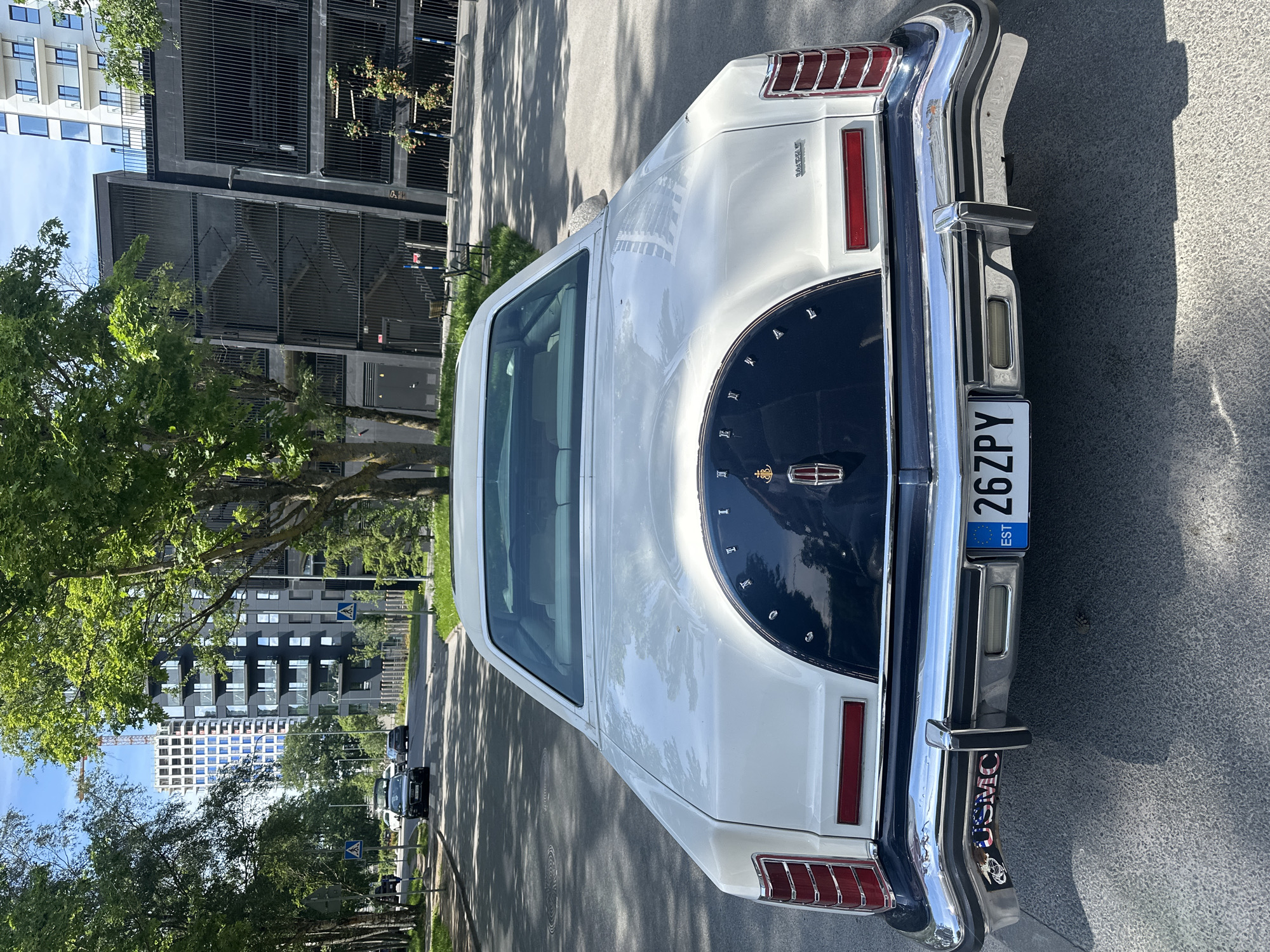 The image shows the rear of a white Lincoln Continental Mark V parked on a paved road in Tallinn, Estonia. The car has a dark blue landau roof section with the Lincoln emblem in the center. The license plate reads "26 ZPY EST". A sticker with "USMC" is visible on the lower left corner of the bumper. In the background, there are modern apartment buildings, trees, and a multi-story parking garage. The sky is blue with some clouds.