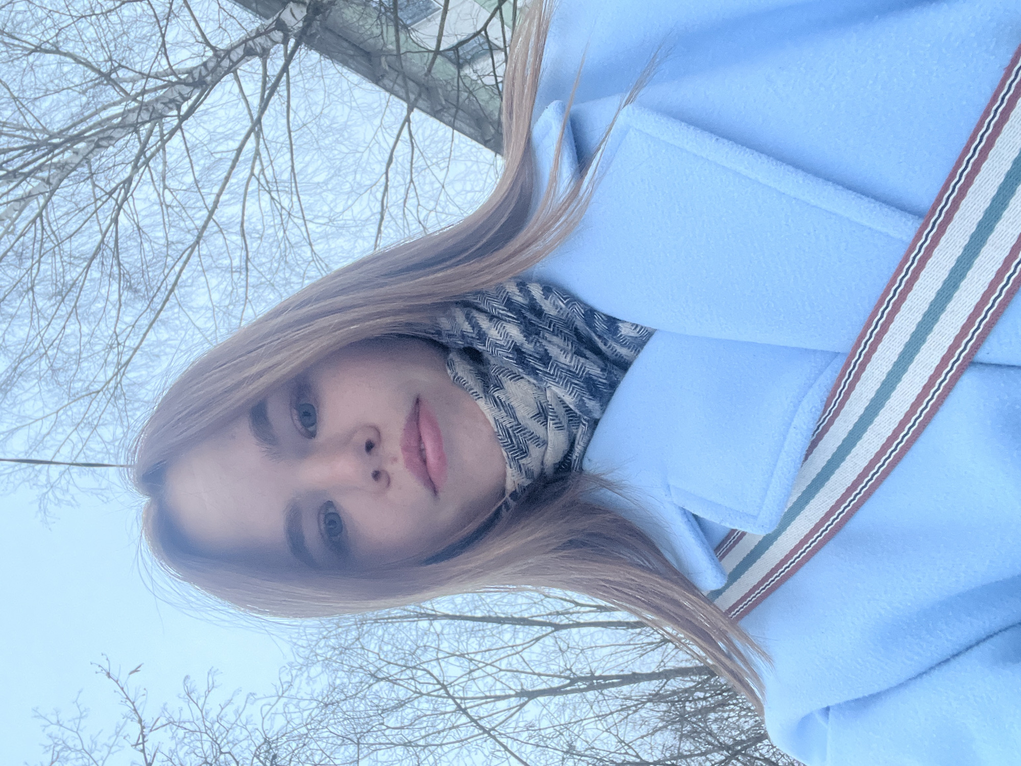 An upward-angled selfie captures a young adult female outdoors in Chaikovsky, Russia. The subject has light brown hair, blue eyes, and a neutral facial expression with slightly parted lips, looking directly into the camera. She is wearing a light blue textured overcoat and a patterned scarf with white, blue, and black geometric designs around her neck. A cross-body strap, featuring white, red-brown, and pale green stripes, is visible over her left shoulder and torso. The background consists of numerous leafless deciduous trees, some with visible lighter bark patterns suggesting birch, silhouetted against an overcast, light-colored sky. A portion of a light-colored building with dark window outlines is visible on the upper right. The environment suggests a late autumn or winter setting.