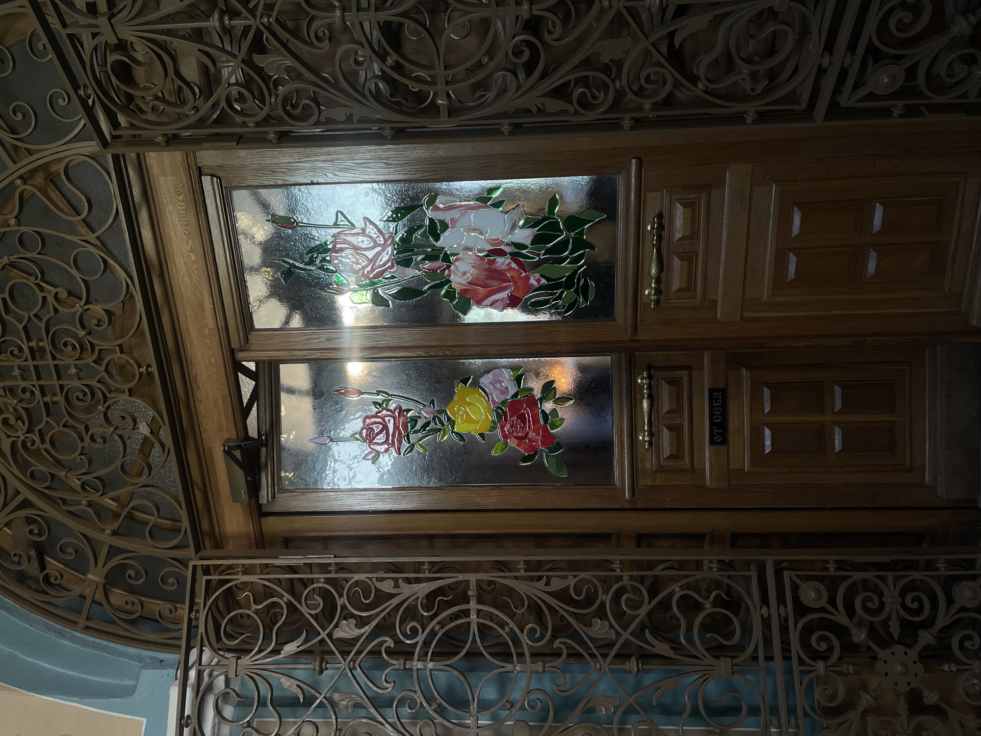 Here is a description of the image:
The image captures an interior view, likely a decorative element within a building in Moscow, Russia. The focal point consists of ornate wooden cabinetry, featuring two glazed panels with a stained-glass floral design. Each panel showcases a bouquet of roses, with the upper panel displaying predominantly white and red roses, while the lower panel includes red, yellow, and pink roses. The glass panels appear textured, giving a slightly blurred or antiqued effect to the floral paintings.
Adjacent to the cabinet doors are decorative metal grilles with intricate scrollwork and floral motifs. The upper portion of the image shows additional decorative metalwork, possibly a ceiling feature. Below the cabinetry, another metal grille is visible, partially obscuring a muted blue wall in the background. A small rectangular sign with lettering in a Cyrillic script is attached to the right cabinet. The scene appears to be illuminated by a warm, natural light, casting subtle reflections on the glass and highlighting the details of the wood and metal.