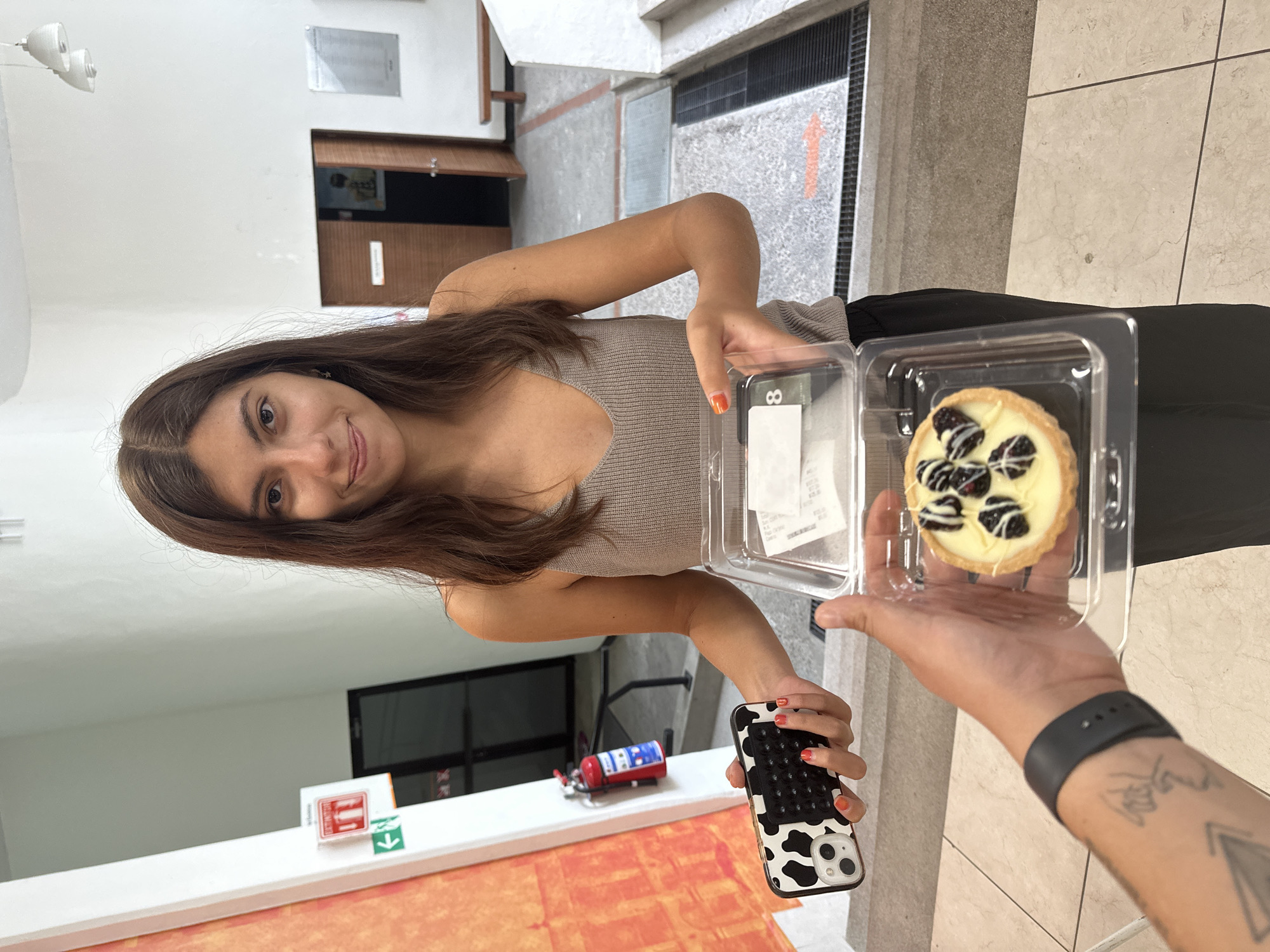 This vertically oriented image captures a moment in an indoor setting, likely a hallway or lobby in San Andrés Cholula, Mexico, during the daytime. The primary subject is a young woman with long brown hair, wearing a light tan tank top, who is looking towards the camera and smiling. She holds a smartphone with a black and white cow-print case and a black pop-it style grip in her left hand, her fingernails painted orange.
To the right, an arm and hand belonging to another person extend into the frame, holding a clear plastic container. Inside the container is a small tart featuring a white cream base topped with several dark berries, possibly blackberries or blueberries, and a decorative white drizzle. A sticker on the container displays the number "8" and a fragment of a receipt showing "B $110.00". The arm wears a black wristband or watch and has visible tattoos, including a geometric design.
The background reveals simple institutional architecture: a white wall on the left, and a grey tiled floor and wall to the right. A red fire extinguisher with a red, white, and blue label is visible on a low white ledge, next to a white sign featuring a red 'I' symbol and a green downward-pointing arrow. Further back, there are dark framed openings, wooden paneling, and a general impression of an administrative or public building. An orange arrow is painted on the grey floor tiles. The woman's pleasant expression suggests a friendly interaction.