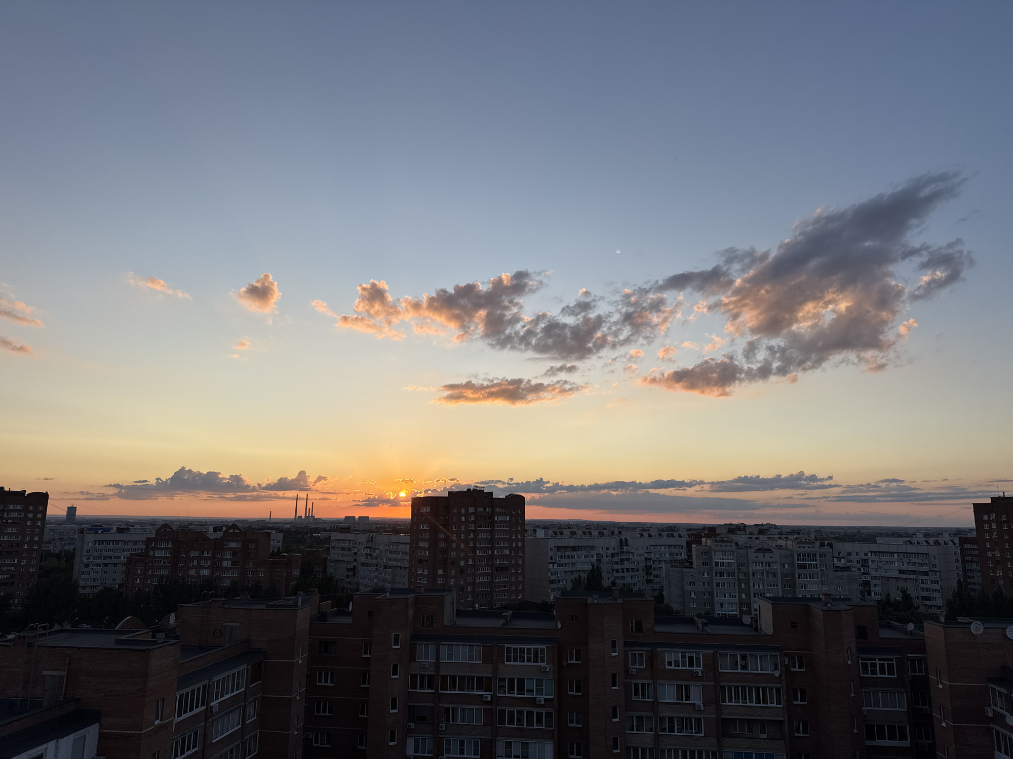 The image shows a cityscape at sunset. The sky is a gradient of blue and orange, with clouds illuminated by the setting sun. The sun itself is partially visible, casting a warm glow over the scene. Numerous buildings, primarily apartment blocks, fill the midground and foreground. They are of various heights and architectural styles, with many featuring red brick exteriors. Some buildings have visible windows and balconies. In the distance, several tall, industrial-looking structures, possibly chimneys or towers, are visible. The overall atmosphere is one of a tranquil evening in an urban environment. The time of day is clearly sunset, suggested by the colors in the sky and the low angle of the sun. There is no visible text in the image.