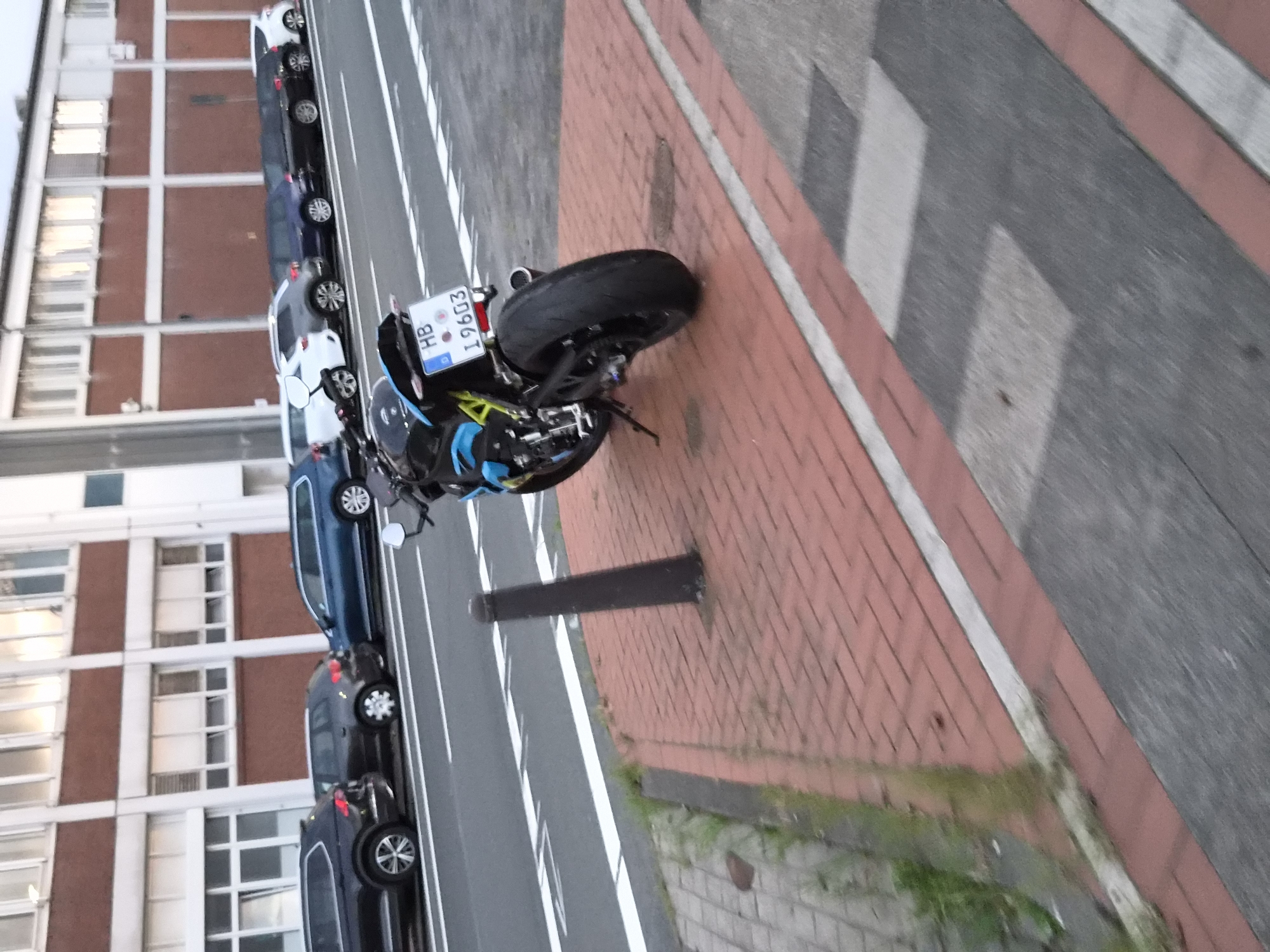 The image shows a street scene in Bremerhaven, Germany. A black and blue motorcycle is parked on a red brick sidewalk. The motorcycle has a license plate that reads "HB I 9603". In the background, several cars are parked along the street in front of a building with white and red brick accents. The street is paved and there are white lane markings. The overall environment appears to be an urban setting during daylight hours.