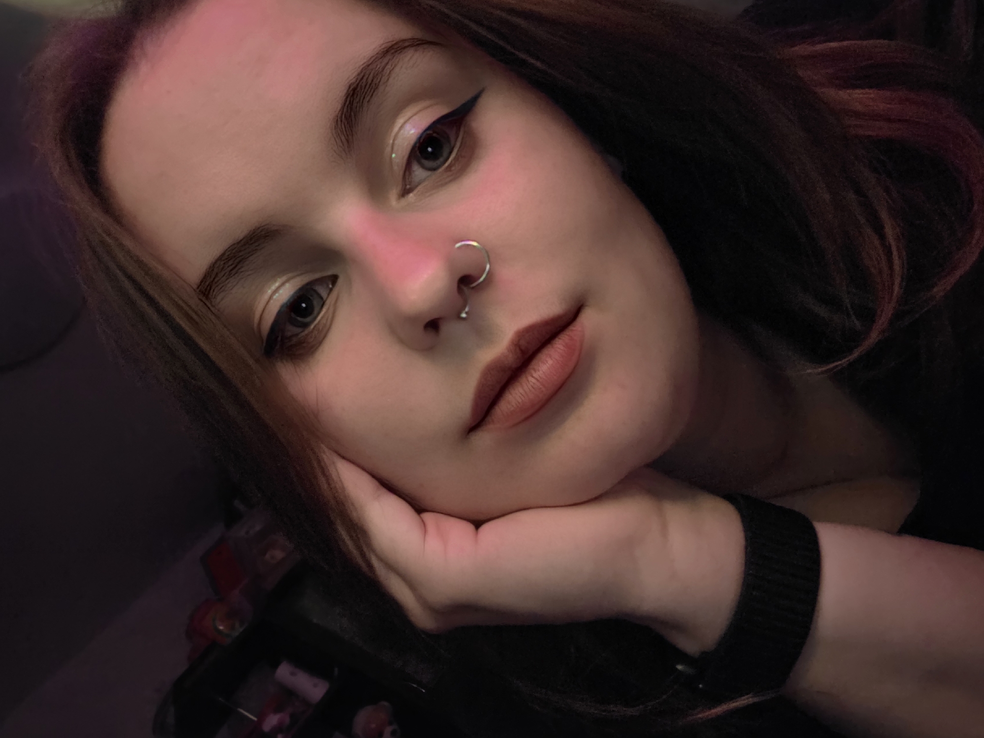 A close-up, high-angle selfie shows a young person with light skin, dark brown hair with auburn highlights, and blue-gray eyes. The person is wearing subtle makeup, including winged eyeliner and lip gloss. A silver septum ring is visible in their nose. Their right hand rests on their cheek, and a black fitness tracker is worn on their left wrist. The background is dark and out of focus, with hints of shelves and various objects, suggesting an indoor setting. The lighting is dim, with a pinkish hue cast over the scene. The image appears to have been taken in a private residence.