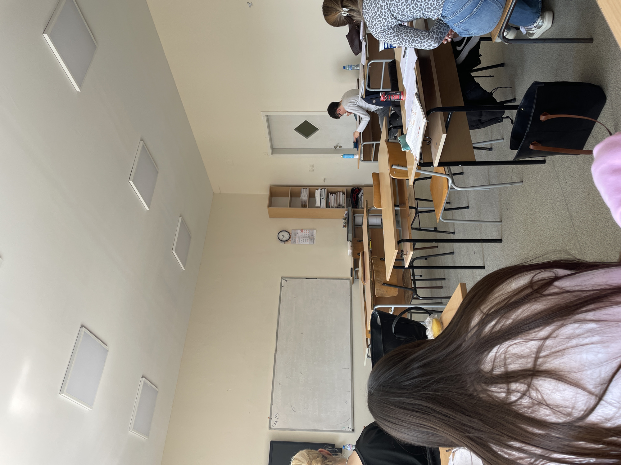 The image depicts an indoor classroom setting. There are multiple rows of student desks and chairs, some occupied by individuals. A whiteboard is mounted on the wall, and a bookcase is visible in the background. The room is illuminated by square ceiling lights. The location is Unknown, Unknown.