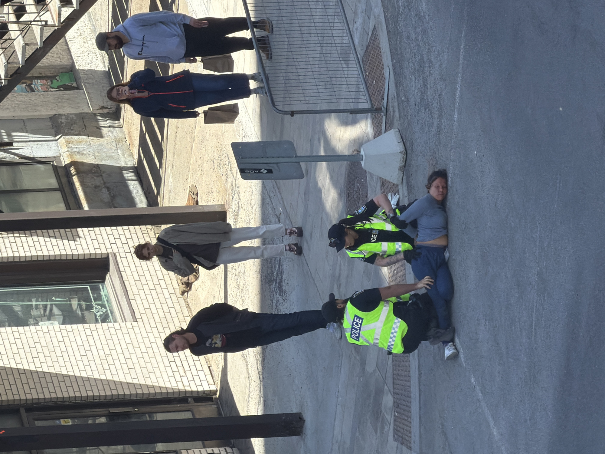 The image depicts an outdoor urban scene featuring a police interaction on a sunlit sidewalk. Two police officers are attending to an individual who is on the ground. One officer is kneeling beside the person, while the other stands nearby. Several bystanders are observing the event from various points on the sidewalk.
The two police officers are distinctly dressed in bright neon yellow-green vests with "POLICE" clearly visible on the back, black pants, and black caps. One officer has tattoos visible on their arm. The person on the ground appears to be a woman, wearing a light blue top, dark blue pants, and white sneakers; her posture and facial expression suggest discomfort or distress. Among the bystanders, a man in a black t-shirt and dark pants stands closest to the activity, looking on. Further away, another individual in a light grey jacket and light pants observes, as does a woman in a dark blue and red jacket. A man in a light grey "Champion" brand hoodie is also present further back.
The setting is a city street, likely in Montreal as per context, characterized by multi-story brick buildings with windows. The strong shadows cast on the pavement and buildings indicate bright sunlight, suggesting a clear day, possibly late morning or early afternoon. Visible objects include a metal crowd control barrier, a street sign with a concrete base (displaying "ADS" in reverse), and several brown paper bags on the ground. A metal railing is visible in the upper left, potentially part of a building's exterior structure. The ground consists of grey paved sidewalk and street surfaces.