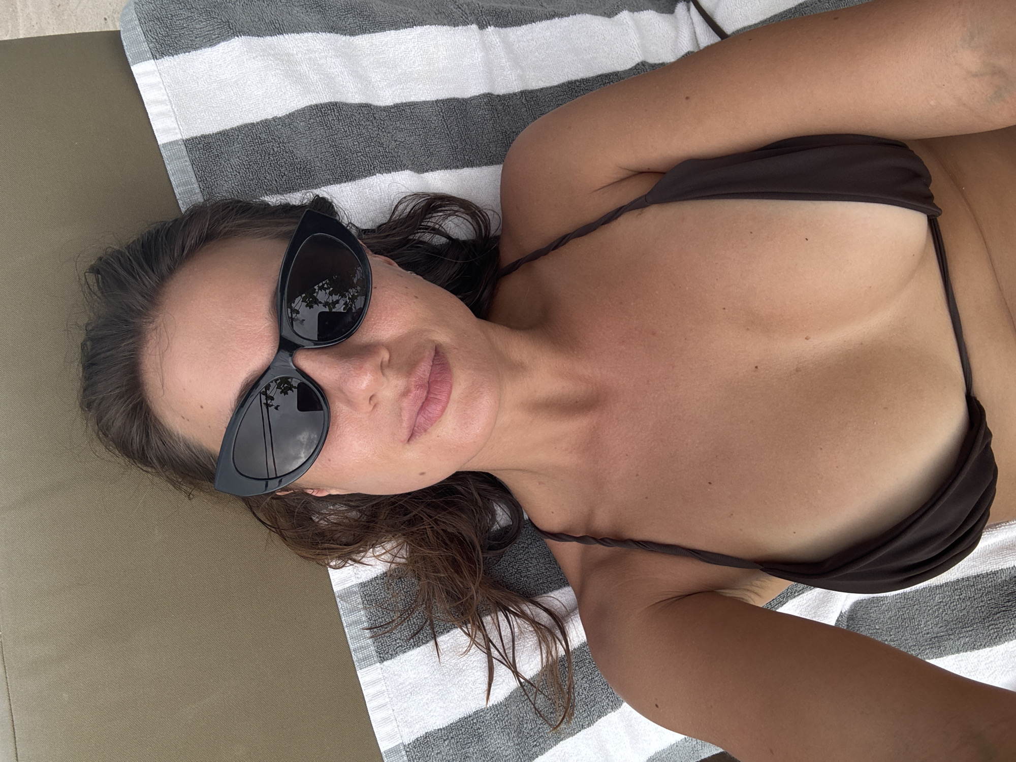 A tanned woman wearing a dark bikini and black sunglasses lies on a gray and white striped towel on a light brown surface. Her hair is brown and damp. The location is Unknown, Indonesia.