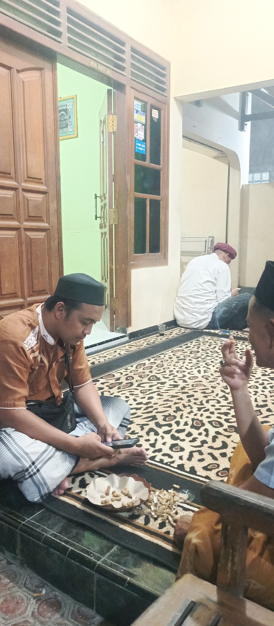 A medium shot captures three men in what appears to be a residential setting in Kedung Dowo, Indonesia. The first man, foreground left, is seated cross-legged on a carpet, wearing a brown, patterned shirt and a black cap. He is looking down at his mobile phone, which he holds in his right hand. Beside him, on the carpet, is a plate containing a small pile of snacks, likely peanuts, with some scattered around it.
To the right of the foreground man, another man is partially visible, seated and facing away from the camera, his right hand raised. He wears a light-colored shirt.
In the background, a third man is seated against a wall, wearing a white shirt and a red cap. He is also looking down, potentially at a mobile phone. A wooden door with a carved panel is visible on the left, with a framed picture hanging on the wall inside the room. The floor is covered with a patterned carpet. The overall environment suggests a casual gathering or a moment of respite.