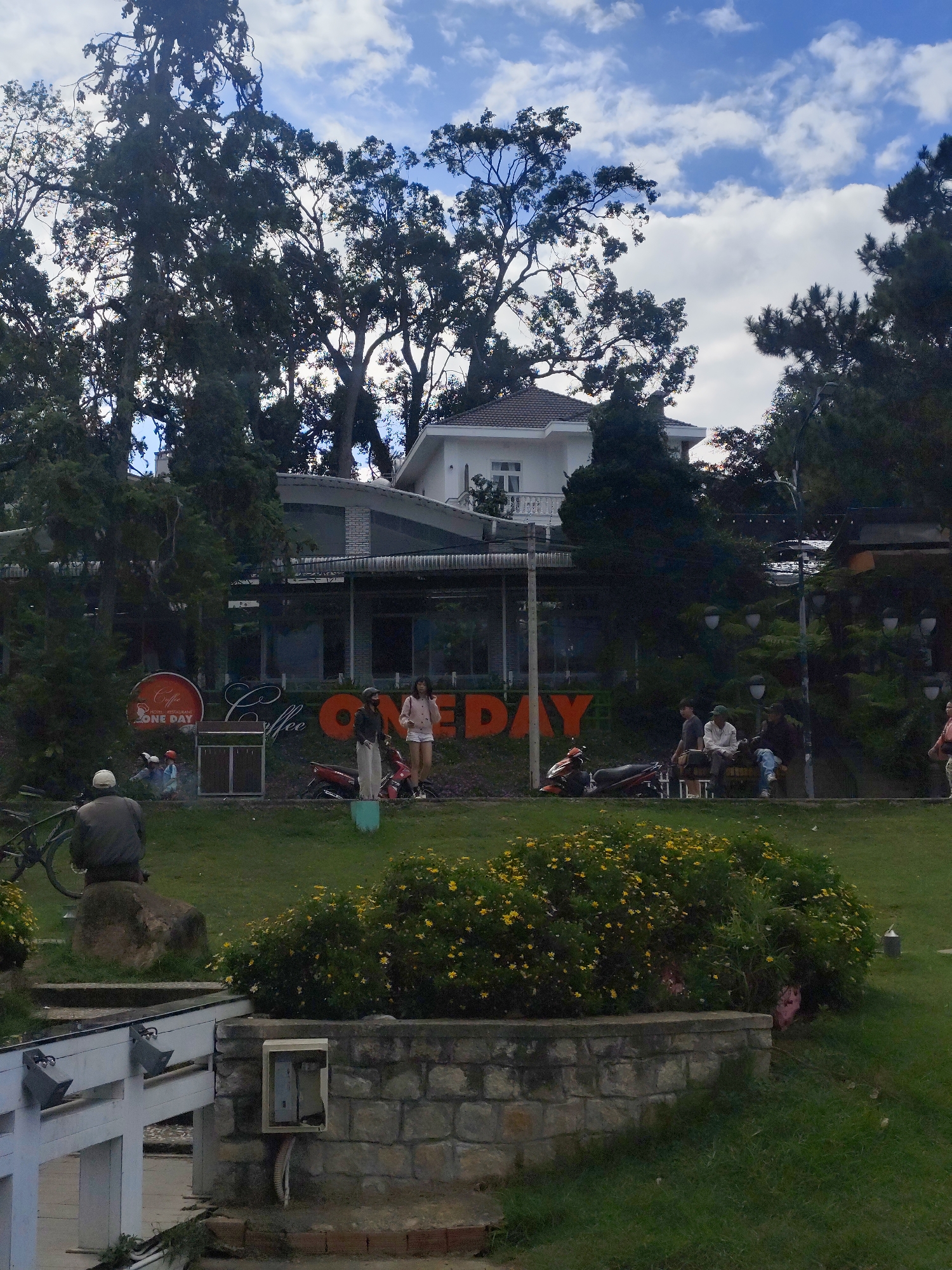 A low-angle, full shot captures an outdoor scene in Da Lat, Vietnam, featuring a cafe called "ONE DAY" with a circular orange sign and large orange lettering spelling "ONE DAY" on its exterior. The cafe is situated in front of a multi-story building with a white facade and dark-tiled roof, partially obscured by dense trees. In the foreground, a stone wall with a white railing borders a grassy area. A man wearing a white cap and dark jacket is seated on a rock facing away from the camera, with a bicycle beside him. Further back, several people are visible near the cafe, including two women standing on the grass and three men sitting on a bench. Two motorcycles are parked near the cafe entrance. The environment includes lush green grass, a large bush with yellow flowers, and mature trees with dark foliage against a partly cloudy sky.