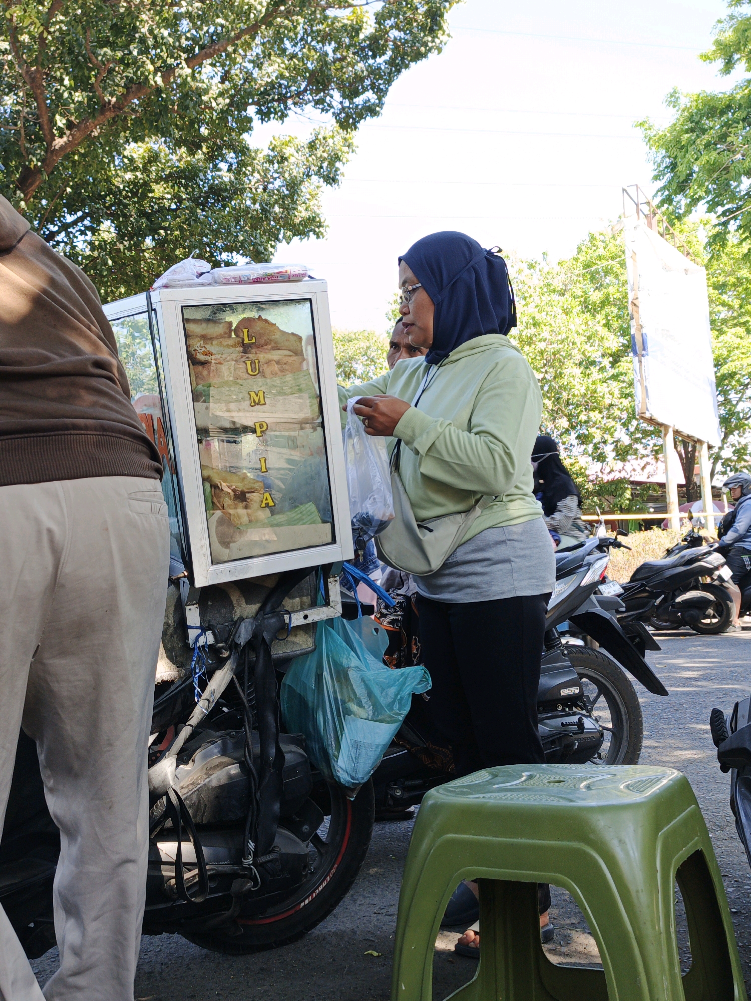 In Sumokali, Indonesia, a woman wearing a dark blue hijab and a light green hoodie is standing next to a food cart attached to a motorcycle. The food cart has a glass display case filled with what appears to be fried food items, and the word "LUMPIA" is visible in yellow lettering on the glass. She is holding a plastic bag, suggesting she is either preparing to serve or has just served a customer. Another person, partially visible on the left, is wearing a brown jacket and light-colored pants. Several motorcycles are parked in the background, indicating a public area, possibly a street or market. The environment is outdoors, with trees and bright sunlight suggesting a warm day. A green plastic stool is in the foreground.