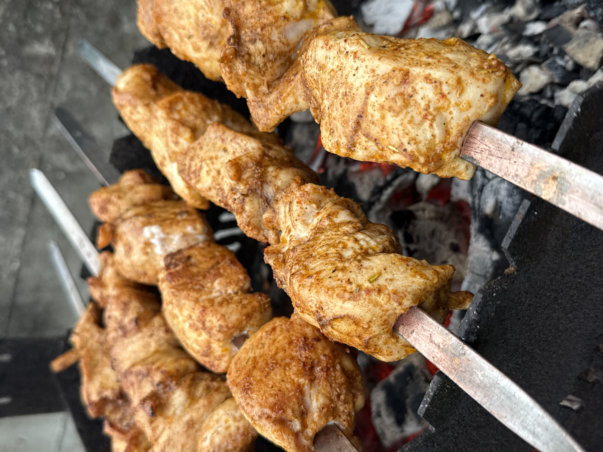 The image shows three metal skewers with grilled chicken pieces, likely shish kebab, cooking over a charcoal grill. The chicken pieces are golden brown with grill marks and appear to be well-cooked. The charcoal underneath is glowing red, indicating high heat. The skewers are positioned on the grill, ready for cooking. The environment is a barbecue setting, possibly outdoors. There are no people visible in the image. The location is unknown.
