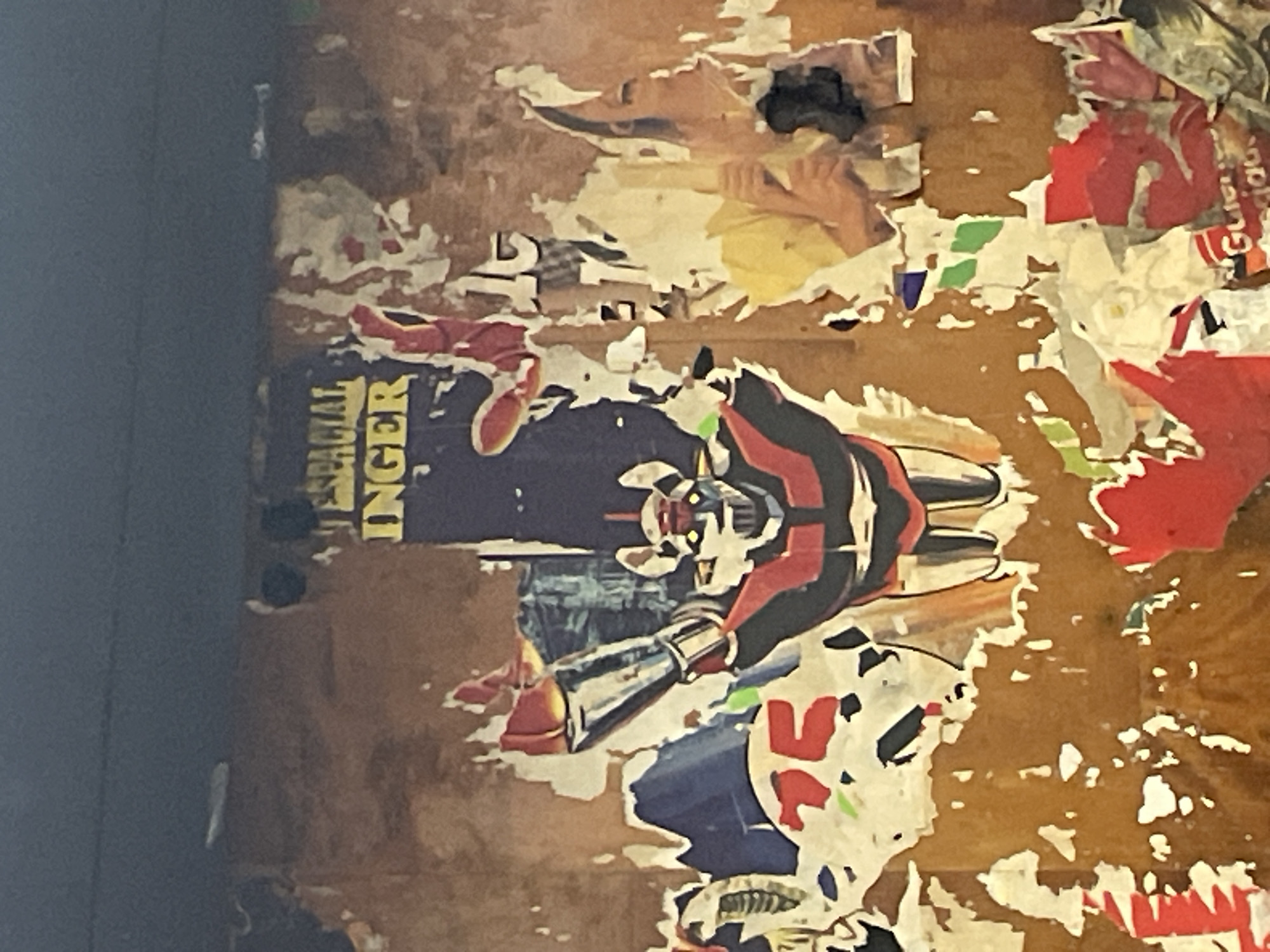 A close-up, slightly angled photograph depicts a section of a bulletin board or wall, heavily layered with torn and decaying posters. The surface is brown and textured, likely wood or a similar material. Dominating the visible section is a partly torn poster of "METAL INGER," a Japanese anime robot character, colored in black, red, and white with a silver cannon arm. Other torn posters with fragments of images and text are visible around the robot poster. This image likely originates from Barcelona, Spain, evidenced by the recognizable poster content commonly found in urban spaces there.