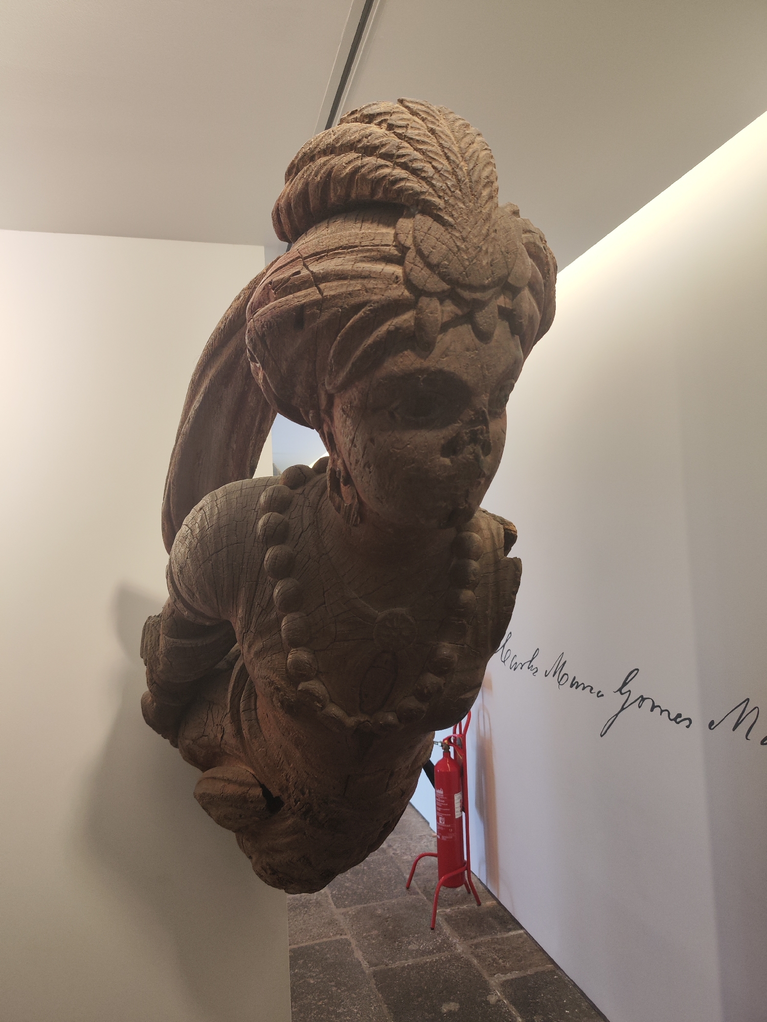 The image displays a large, intricately carved wooden figure, likely a bust or a figurehead, mounted on a pristine white wall in an indoor exhibition space. The sculpture depicts the upper body of a person, possibly female, adorned with an elaborate turban or headwear featuring a prominent feather-like ornament. The figure's face has stylized or weathered features, and it wears a beaded necklace and decorative elements on its torso. The wood is a rich, dark brown, showing signs of age with visible cracks and texture.
The figure is positioned at an angle, creating a dynamic visual. The surrounding environment consists of stark white walls and a light-colored ceiling, with integrated lighting illuminating the space. The floor is dark, appearing to be made of stone or large tiles. To the right of the wooden figure, a bright red fire extinguisher stands on a matching red base against the wall. Black cursive text is visible on the white wall, partially obscured, reading "...works Moones Gomes Ma...", suggesting an attribution or title for the exhibit. The setting suggests a museum or gallery in Ponta Delgada, Portugal, dedicated to displaying artifacts or art. No people are visible, and the scene is static, focusing on the displayed artwork.