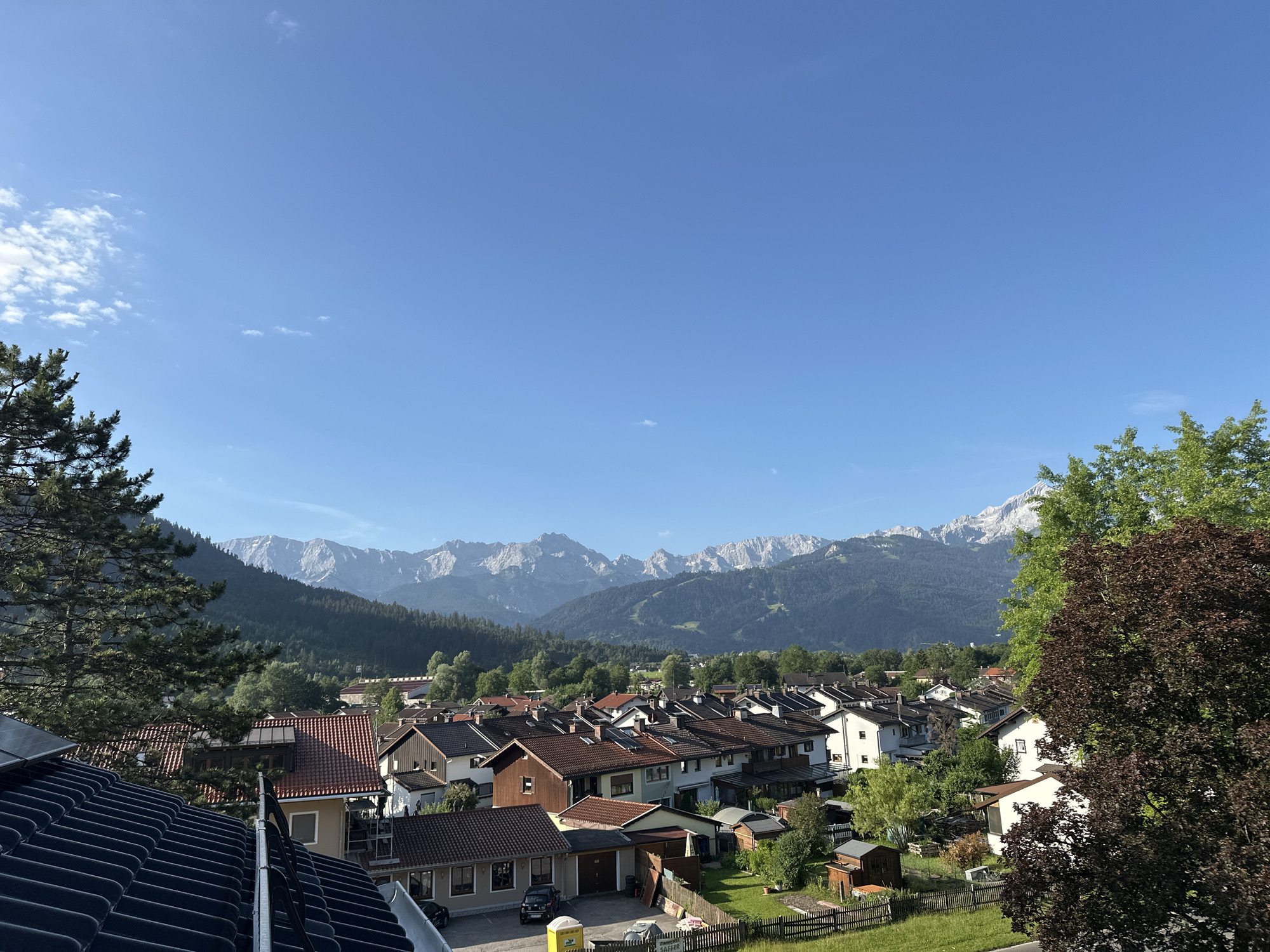 This image presents a high-angle view of a residential area in Garmisch-Partenkirchen, Germany, with a majestic mountain range in the background. The foreground is dominated by the dark, ribbed roof of a building, likely a house, with solar panels visible on the left.
The middle ground features a dense cluster of houses, characterized by their red-tiled roofs and a variety of architectural styles, ranging from traditional Bavarian to more modern designs. Some houses have visible garages, and gardens with fences and small sheds are interspersed between them. A yellow portable toilet is situated in a driveway.
The background is defined by a lush, green forested mountainside that ascends to a dramatic mountain range with snow-capped peaks, indicating a high altitude and possibly summer conditions. The sky is a clear, bright blue with a few wispy clouds. The overall impression is of a peaceful, scenic European town nestled against a stunning natural backdrop. There are no visible people or animals in the scene. The lighting suggests it is daytime, likely morning or late afternoon, given the long shadows.