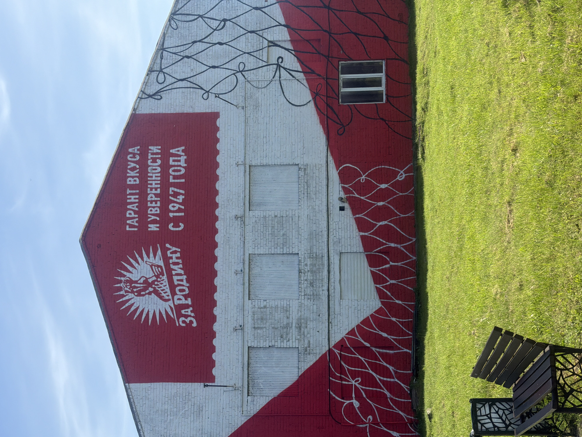 A wide shot captures the side of a brick building, painted white and red, featuring a mural. The mural depicts a stylized sunburst logo with the words "ЗА РОДИНУ" (For the Motherland) in white, and to its right, "ГАРАНТ ВКУСА И УВЕРЕННОСТИ С 1947 ГОДА" (Guarantee of Taste and Confidence Since 1947) in white lettering against a red background. The building has several windows, some covered with ribbed panels and one with a visible pane, all painted white. A large red diagonal stripe runs across the lower portion of the building, decorated with a white, net-like pattern. A black net-like pattern is also present on the upper right section of the building. The building is situated in a grassy field, with a dark wooden bench and a decorative black post in the foreground. The sky above is blue with scattered white clouds. The location is identified as Vzmorye, Russia.