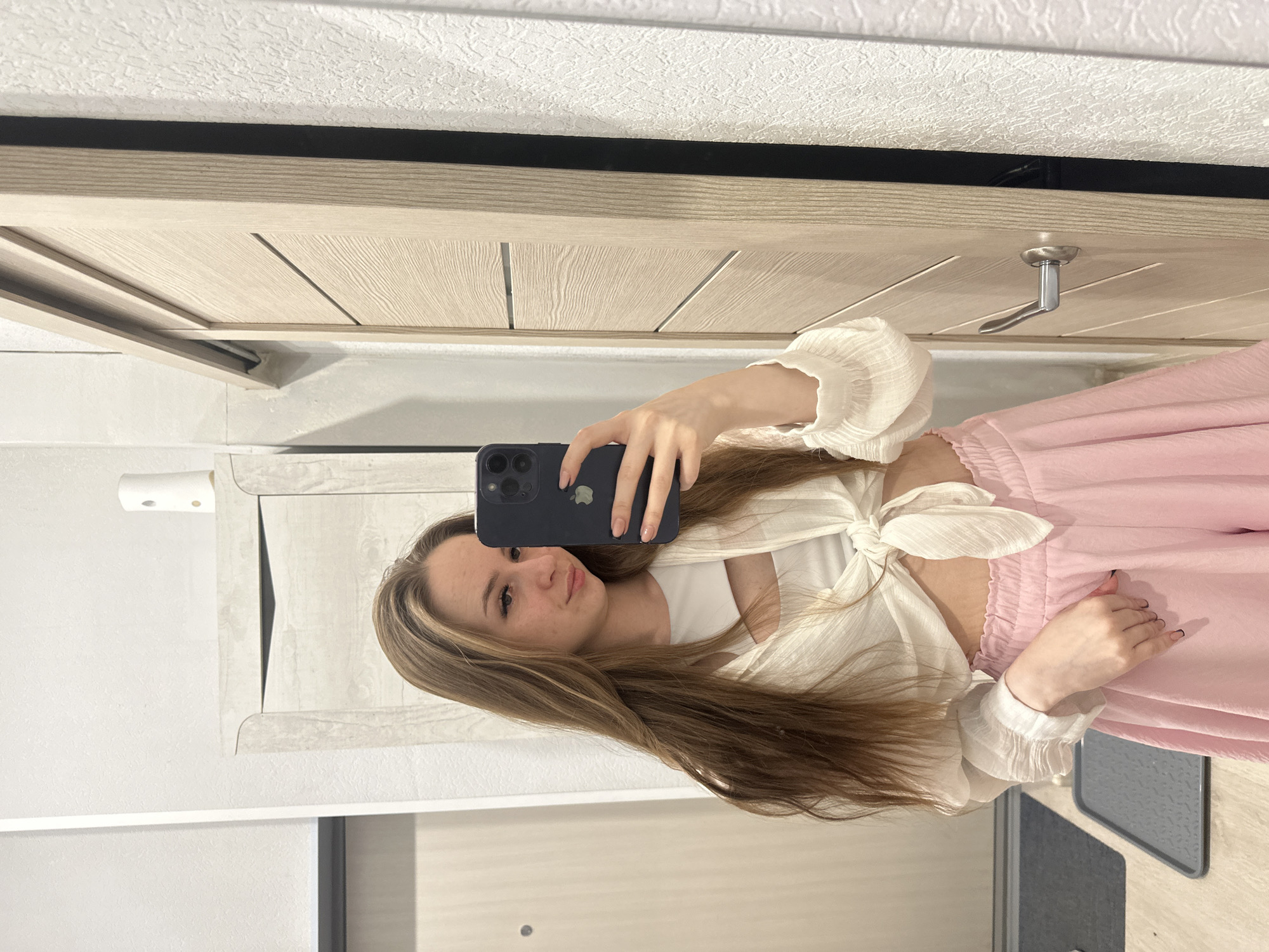 A young woman is taking a selfie in what appears to be a bathroom setting. She is holding a dark-colored smartphone, likely an iPhone, up to her face, obscuring the upper portion of her face. She has long, brown hair with blonde streaks. She is wearing a white crop top with a white, tied-front blouse and pink sweatpants. The background shows a mirrored surface. A portion of a cabinet or door is visible above her, with a silver door handle. The overall environment appears to be a bathroom setting, with white or light-colored walls and fixtures. The specific location in the city of Unknown, Unknown cannot be determined from the image.