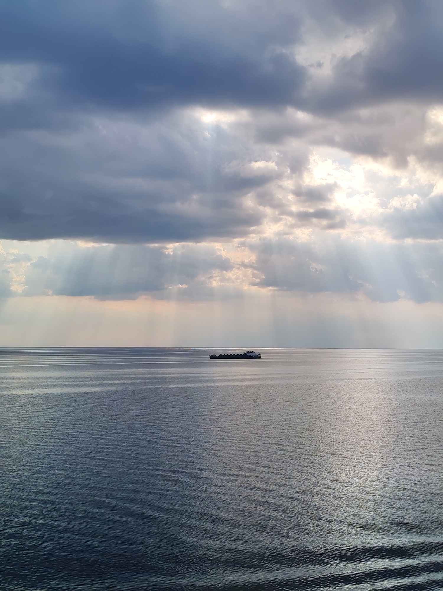 The image depicts a calm body of water, likely a lake or wide river, under a sky filled with dramatic clouds. Sunbeams are piercing through the clouds, creating a contrast between light and shadow on the water's surface. A long barge or vessel is positioned on the horizon. Ripples are visible near the bottom of the image, suggesting the viewpoint is close to the water's edge.
While it is implied that the location is Belozersk, Russia, there are no elements in the image that concretely confirm that the location is Belozersk. Therefore, the location is inferred.