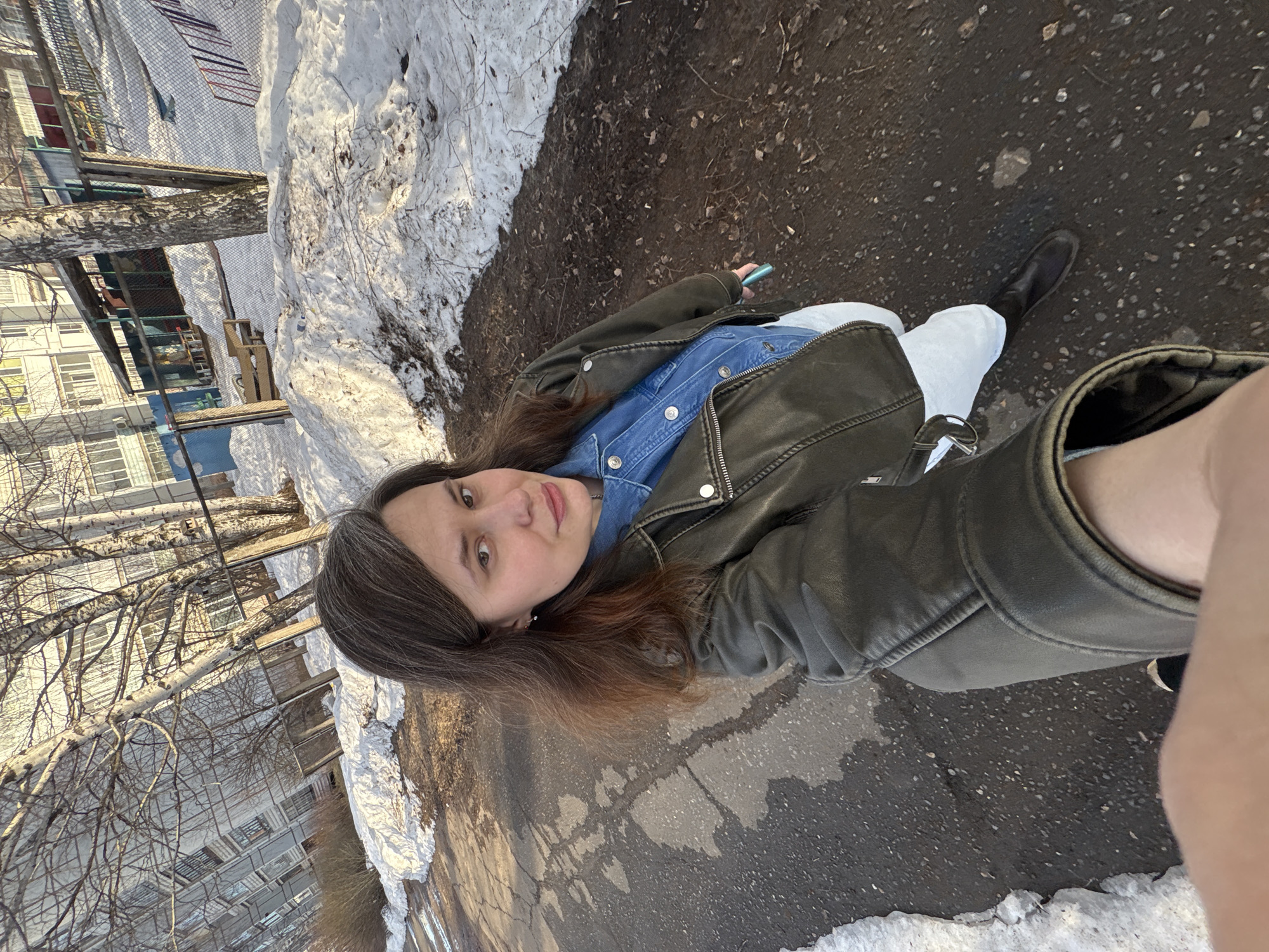 A woman with dark brown hair, interspersed with visible gray strands, is depicted taking a selfie outdoors in the city of Chaikovsky, Russia. She wears a dark olive green leather-style jacket over a blue denim shirt, light-colored trousers, and black boots, and holds a turquoise-colored mobile phone in her right hand. Her expression is neutral as she looks directly at the camera. She stands on a cracked, wet asphalt surface, bordered by irregular patches of melting snow and exposed earth. The background features several bare deciduous trees, identifiable as birches by their light bark, positioned in front of multi-story, light-colored residential apartment buildings. A wire mesh fence runs through the middle ground, partially obscuring a snow-covered area with scattered wooden structures and colorful elements, indicative of a playground. The scene is illuminated by natural daylight, suggesting late winter or early spring conditions.