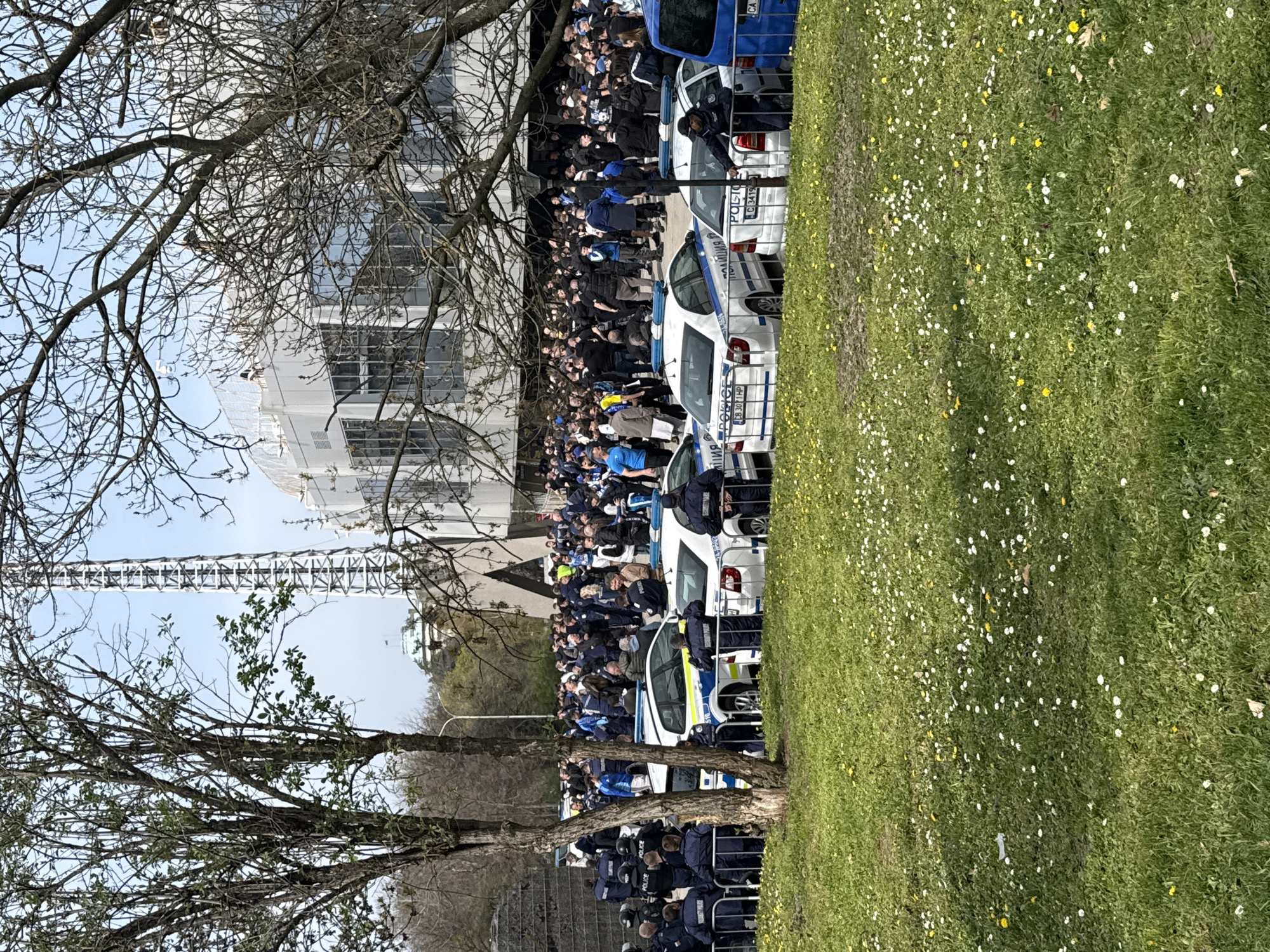 The image shows a crowd of people gathered behind a line of police cars in what appears to be a public area. The location is Unknown, Unknown.
The crowd includes people of varying dress, some wearing blue clothing, suggesting team colors. A line of police officers stands behind the police cars, facing the crowd. The cars are white with blue markings and are marked with "ПОЛИЦИЯ" in Cyrillic script, suggesting the scene is in Bulgaria.
To the side of the crowd is a grassy field with daisies. Bare trees are in the foreground. In the background is a building with modern architecture and what appears to be a transmission tower.