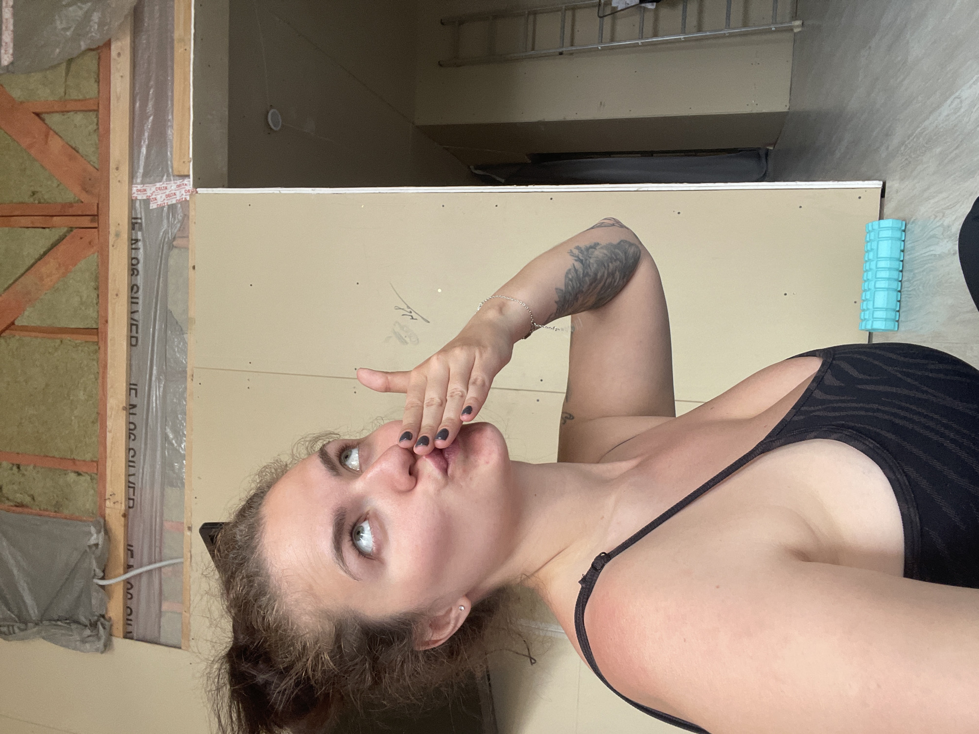 The image depicts a woman with light skin, blue eyes, and dark hair, posing with her hand near her mouth as if blowing a kiss. She is wearing a black top. Her left arm has a tattoo. The background appears to be an unfinished room or a room under construction. There is exposed insulation covered in plastic, wooden beams, and drywall. At the top of the frame, a ladder and a glimpse of another room can be seen. To the right of the woman is a blue textured cylinder, possibly a foam roller. The image appears to have been taken in portrait mode and then rotated. It is not possible to determine the location as Мосметаллоконструкция Онт, Russia from the given image.