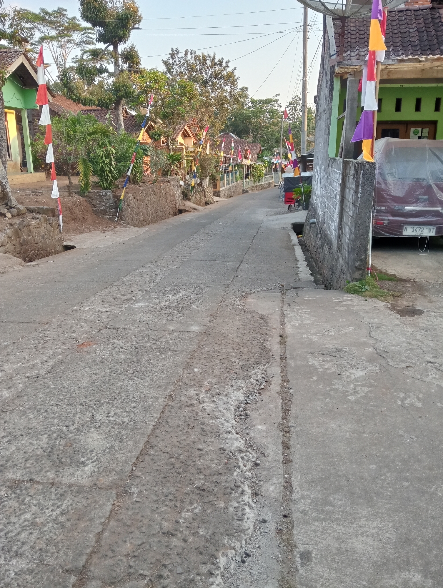 The image depicts a narrow, paved road in Krajan Kidul, Indonesia, lined with houses adorned with colorful flags. The road is cracked and uneven, with a stone wall and a drainage ditch running along the right side. A dark-colored vehicle, possibly a car or van, is parked on the right side of the road, partially covered with a tarp. Several houses with red tile roofs are visible on both sides of the road, some with green walls. Trees and vegetation surround the houses, adding to the rural atmosphere. The sky is clear and bright, indicating a sunny day. The flags, a mix of red, white, purple, and yellow, suggest a festive occasion or cultural event. There are no people visible in the image.
