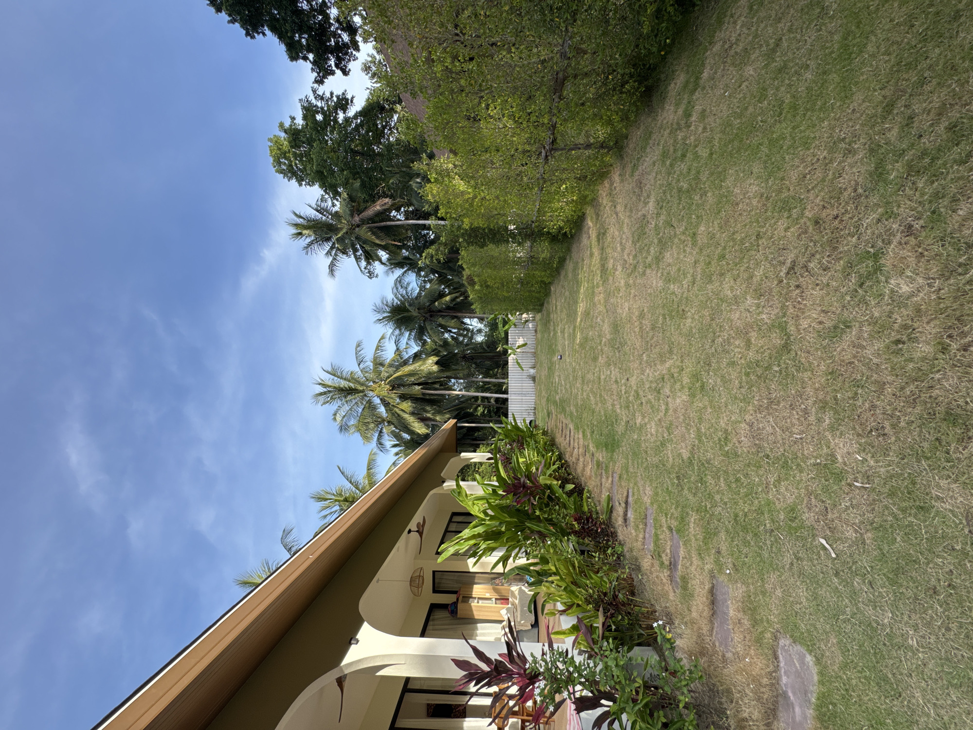 This is a photograph of a house and its surrounding grounds in ตำบลเกาะพะงัน, Thailand. The house has a wide covered porch supported by arched columns, with a brown roofline. In front of the house, there is a garden with lush green plants and some reddish-purple foliage. A path of flat stepping stones leads across a well-maintained lawn towards a white fence.
Beyond the lawn, a dense green hedge runs along the right side of the property. In the background, several tall palm trees are visible, indicating a tropical environment. The sky is blue with scattered white clouds. The overall impression is one of a well-kept tropical residence on a sunny day.