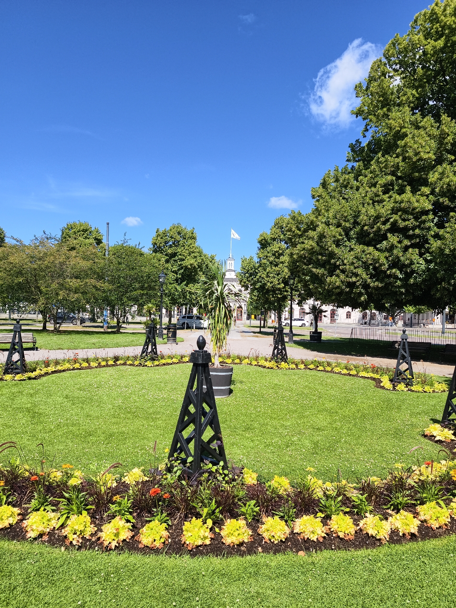 The image depicts a sunny day in a park in Norrköping, Sweden, specifically the area around Rådhuset (City Hall). In the foreground, a circular flowerbed with yellow and orange blooms borders a lush green lawn. Black, lattice-like structures with finials are positioned around the lawn, marking pathways or garden features. Beyond the lawn, several large, mature trees provide shade. In the background, the distinctive white building of Rådhuset is visible with a flag flying from its tower. Several cars are parked along a street that runs behind the park, and a lamppost stands near a trash receptacle. People are not clearly visible or identifiable in the image. The sky is a vibrant blue with a few scattered white clouds. The scene conveys a sense of calm and order in an urban public space.