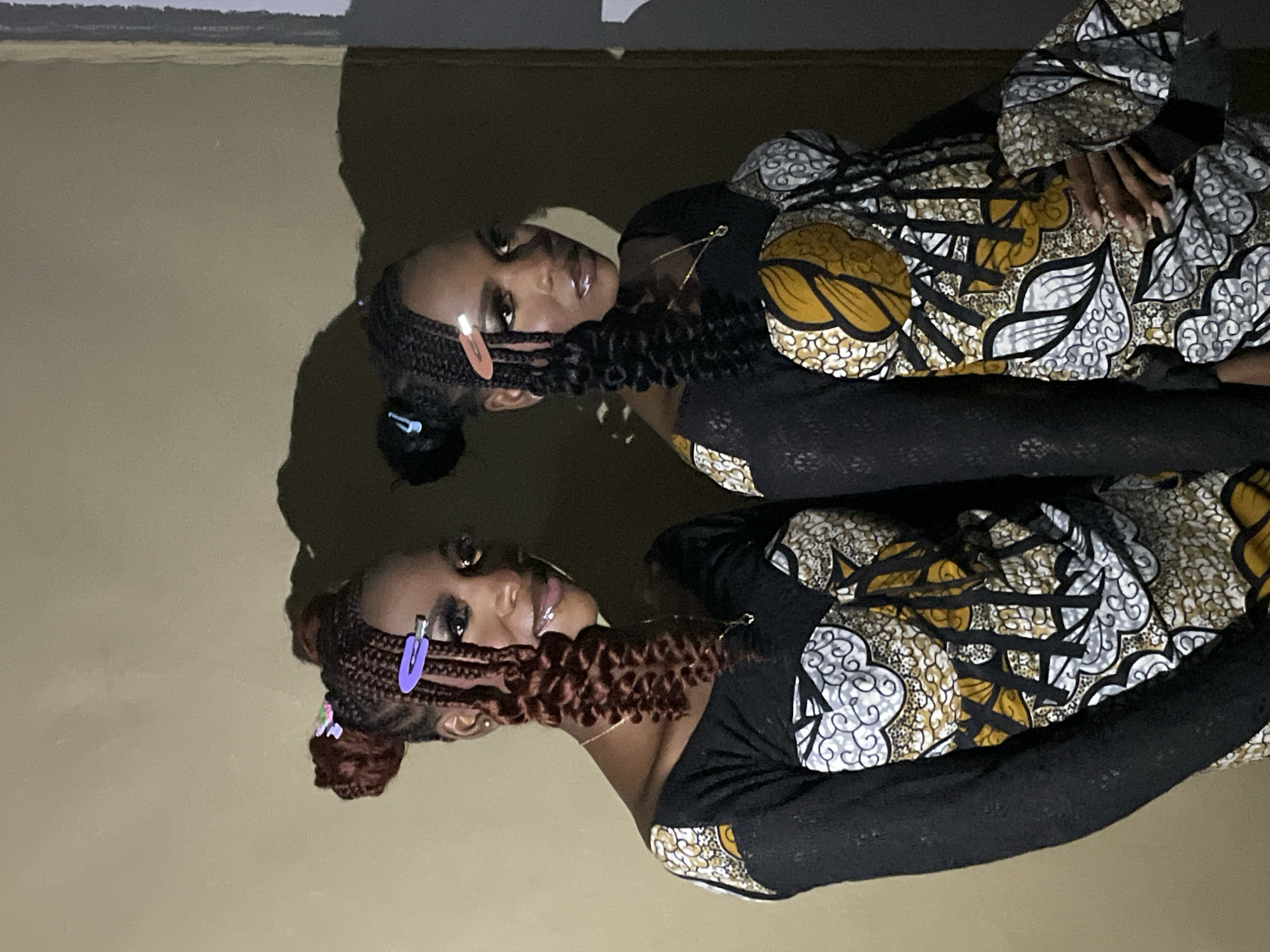 Two young women are posing for a photo indoors. The woman on the left has her head turned to the right, looking at the camera with a slight smile. She has braided hair in two buns, with some braids hanging down her face. The braids are adorned with purple and light pink hair clips. She is wearing a black long-sleeved top with a patterned fabric overlay on the bodice, featuring a yellow, white, and black print with circular and floral motifs. She is also wearing a delicate gold necklace. The woman on the right is partially visible, facing forward and looking slightly to her left. She also has braided hair styled in buns, with braids hanging down. Her braids are adorned with a light blue hair clip. She is wearing a similar outfit to the woman on the left. The background appears to be a plain, light-colored wall with shadows cast upon it, suggesting artificial lighting. The overall impression is that they are in a studio or a room with controlled lighting. This scene is taking place in Durumi, Nigeria.