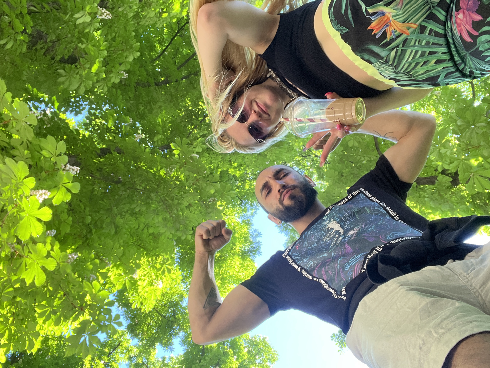 A low-angle shot captures a man and a woman posing in front of a canopy of green trees with glimpses of blue sky. The man, positioned in the foreground, is flexing his arm, showing off a tattoo on his bicep. He has a beard, dark hair, and wears a black t-shirt with graphic print and white shorts. The woman is holding a plastic cup with straws and has long blonde hair and sunglasses. She wears a black top and patterned shorts. The trees are lush and green, forming a natural frame around the subjects. The location is Plovdiv, Bulgaria.