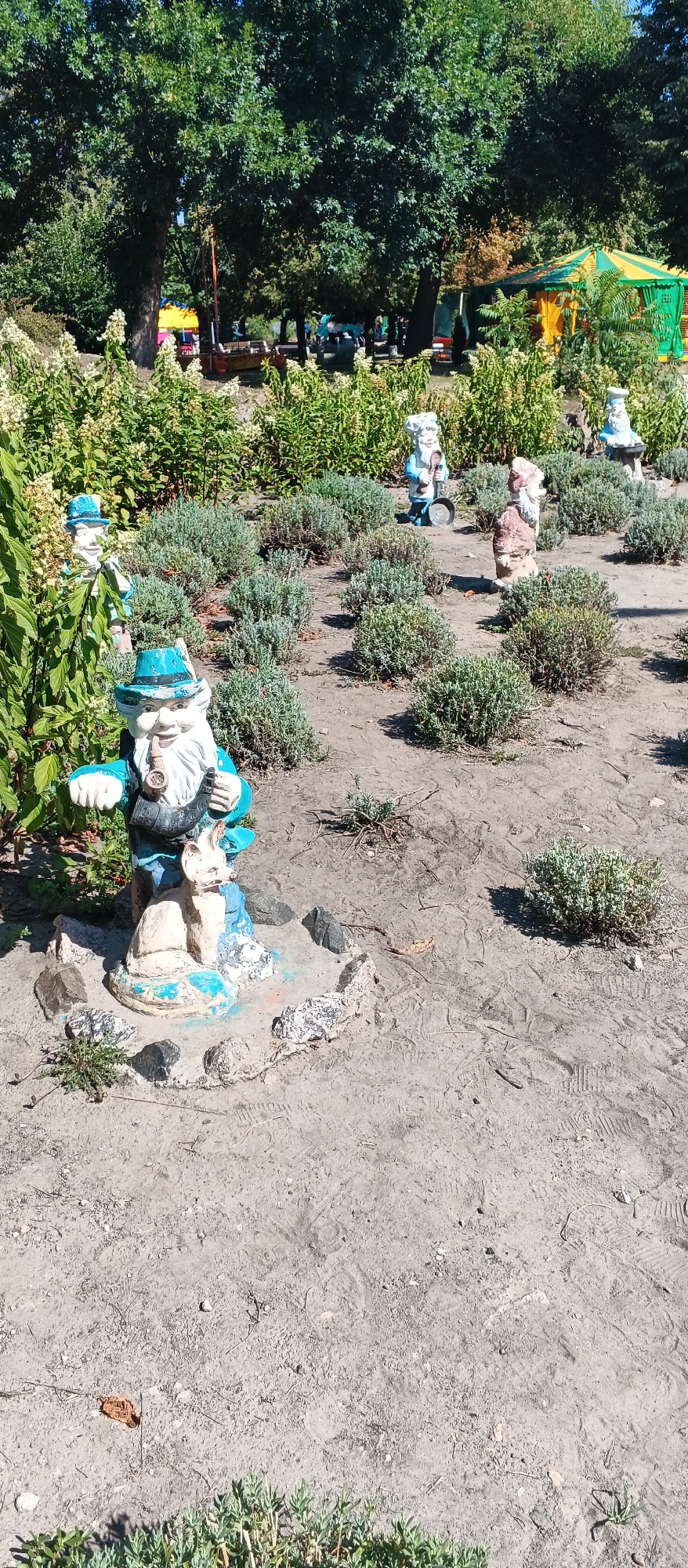 The image shows a garden decoration of gnomes in a park setting. In the foreground, a gnome holding a pipe and a small animal figure are placed on a pedestal surrounded by rocks. In the background, several other gnome statues are visible, some with tools. A large tree with green foliage dominates the left side of the image, with a colorful tent-like structure partially visible behind it on the right. The ground is mostly dirt with scattered stones and patches of low-lying green plants. The scene appears to be in a public outdoor area, likely a park or a backyard in Cherkasy, Ukraine, given the context. The overall impression is of a whimsical and decorative garden.