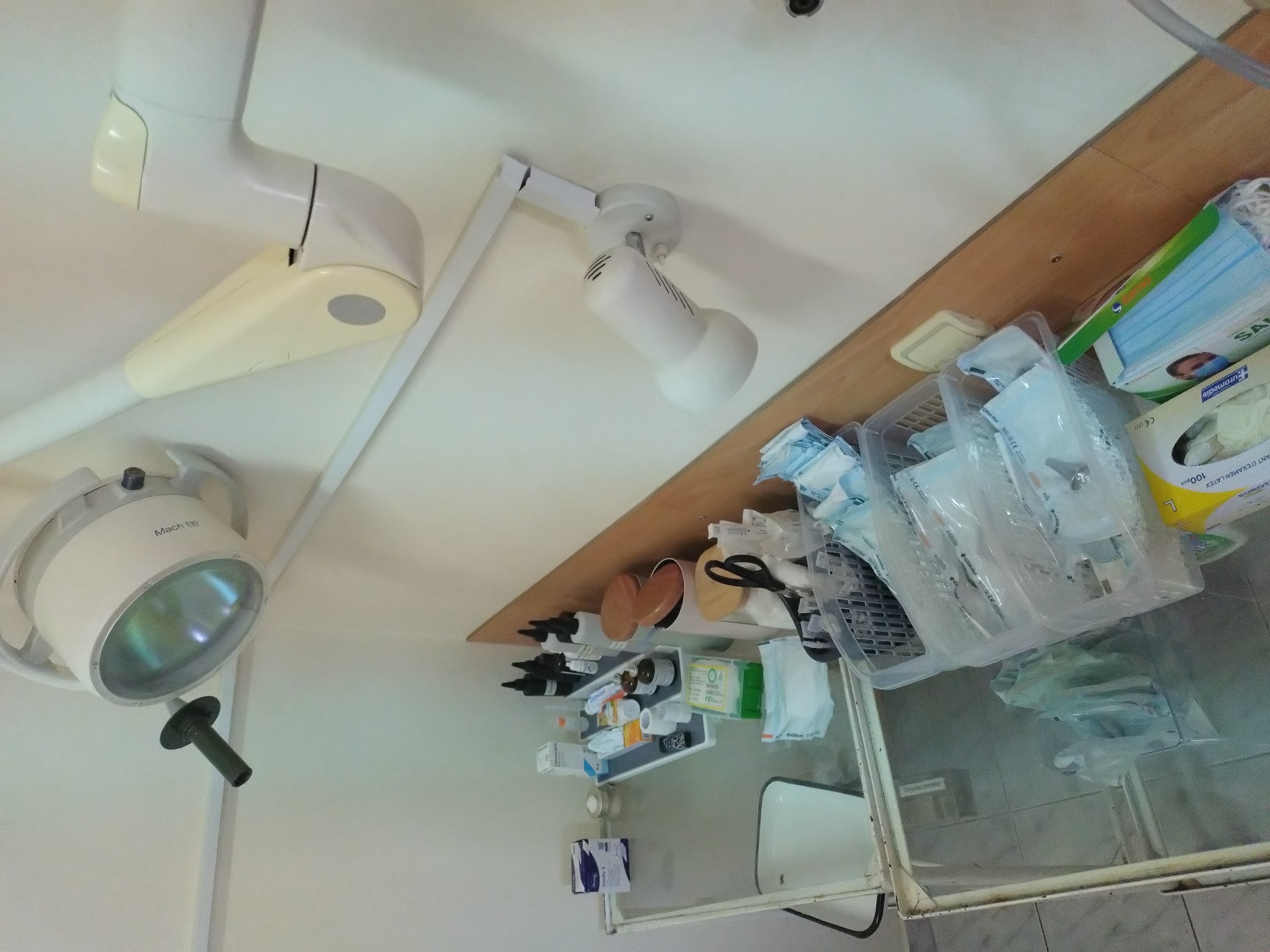 The image presents a clinical setting, likely a medical examination room or a minor surgery area. Overhead, a surgical lamp, labeled "Mach 130," is mounted on a white articulated arm, suggesting its adjustability. Adjacent to this, another smaller, cylindrical white light fixture is also ceiling-mounted.
A wooden shelf runs along the upper part of the wall, holding various medical supplies. These include containers of antiseptics or medications, a pair of scissors, and disposable medical supplies in plastic packaging. Masks in a box are also present. Below the shelf, a smaller shelf unit contains more medical supplies and bottles, implying a structured organization of the materials.
In the foreground, a stainless steel sink is visible, possibly part of a sterilizing or preparation station. The room has a sterile aesthetic, with white walls and a functional design.