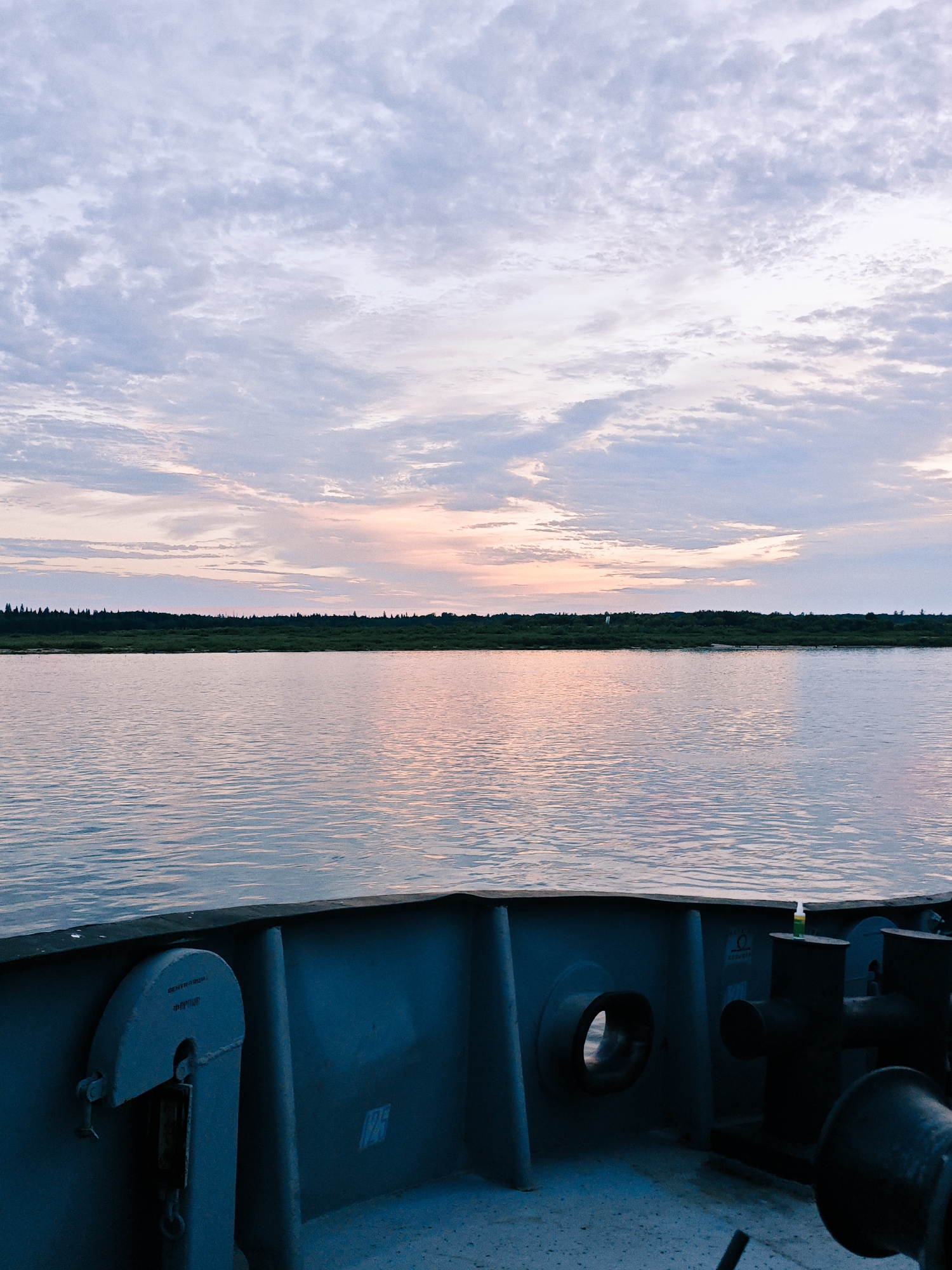 A serene water landscape is depicted, likely from the perspective of a vessel, with the bow of a ship visible in the foreground. The water is calm, reflecting the soft hues of a twilight sky. A distant shoreline is lined with dark, dense vegetation, suggesting a natural, undeveloped area. The sky is a canvas of pale blues and pinks, with scattered clouds, indicative of either dawn or dusk. In the foreground, the metallic structure of the ship's deck is visible, featuring various pieces of equipment, including a capstan and mooring bits. The overall atmosphere is peaceful and quiet, capturing a moment of stillness on the water. The image does not contain any people or any identifiable landmarks that would suggest the location in the city of Nikulino, Russia.