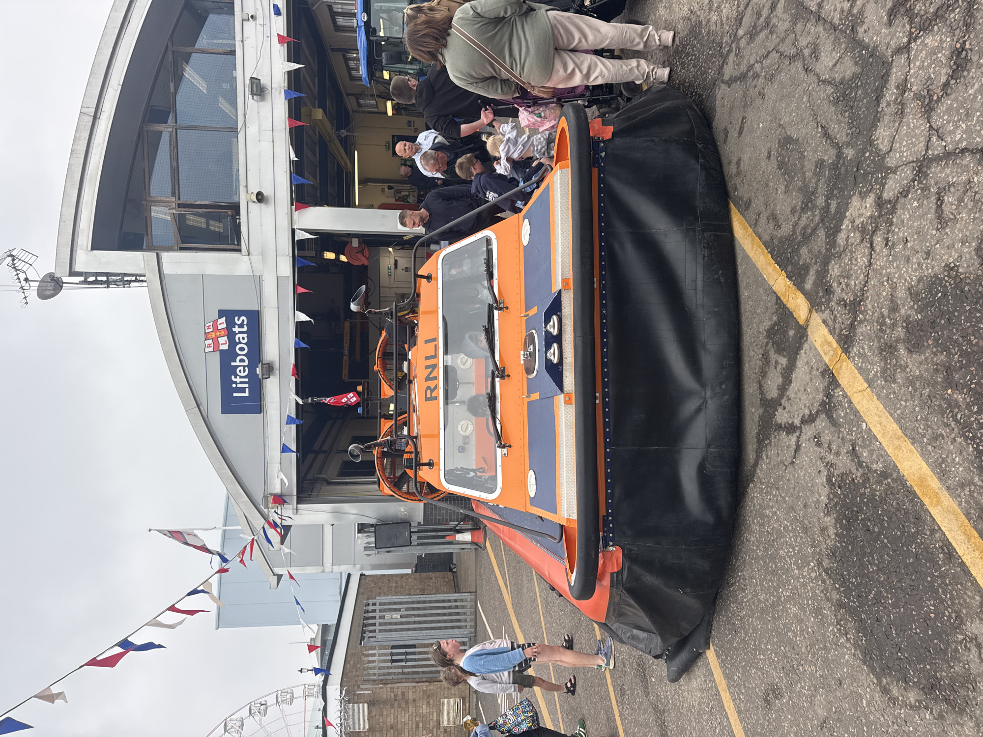 The image shows an RNLI hovercraft on display outside the Southend-on-Sea lifeboat station in the United Kingdom. The hovercraft is orange and blue with a black skirt. Several people are milling around it. Some are looking at the hovercraft, while others appear to be taking photos. The lifeboat station is a modern building with a blue and white sign. There are red, white, and blue bunting flags hanging from the building. The ground is paved with grey asphalt. In the background, a portion of a Ferris wheel is visible.
