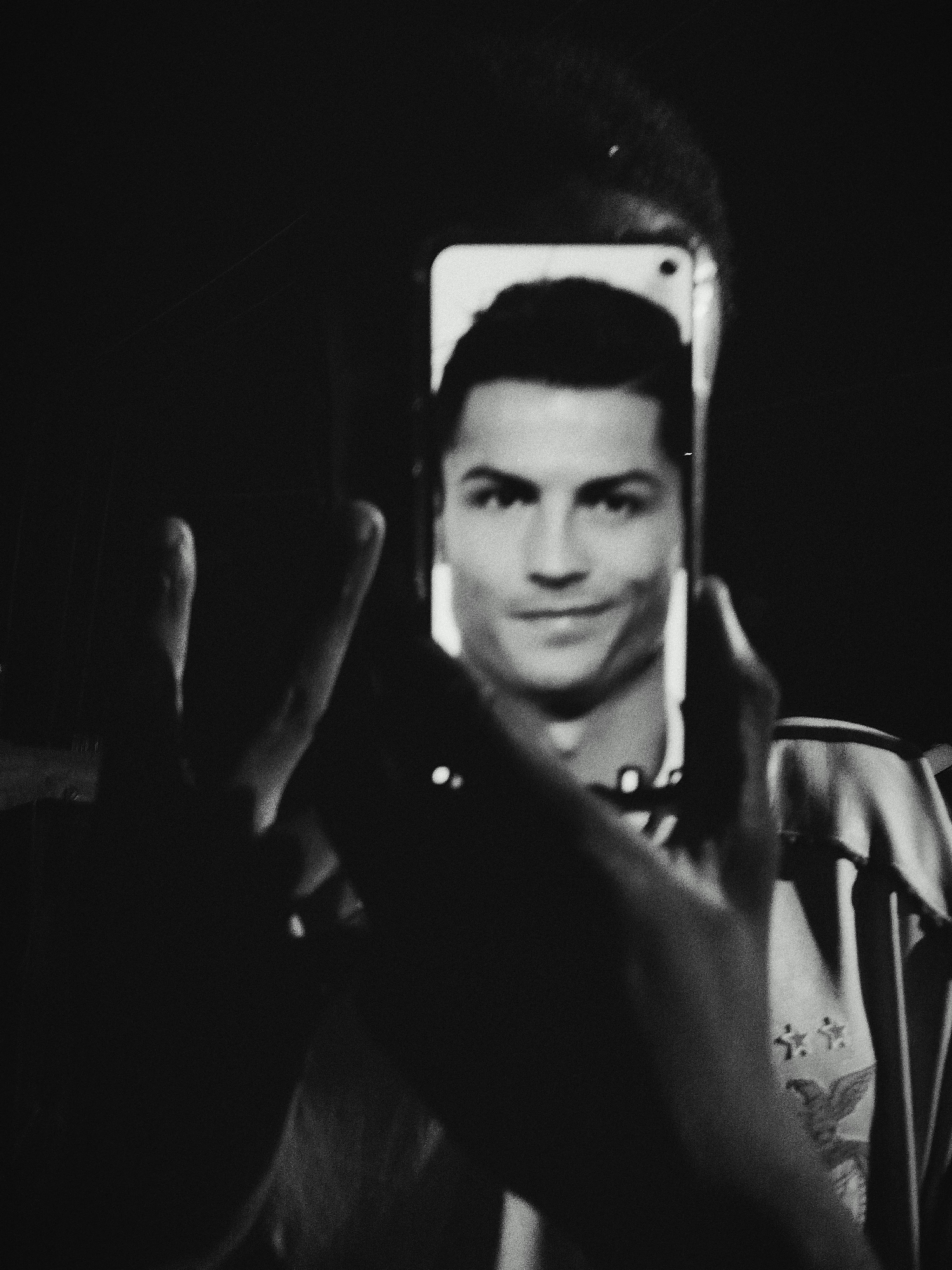 The black and white media file shows a selfie taken by a person holding a smartphone. The smartphone screen displays a portrait of Cristiano Ronaldo. The person taking the selfie is holding the phone in their hands, which are partially visible in the foreground and slightly out of focus. Ronaldo's face is clear and in focus on the screen, showing him smiling. He is wearing a dark top.
The background is dark and indistinct, making it difficult to determine the exact setting or time of day. However, the overall mood appears to be casual and personal, typical of a selfie. There are some faint reflections on the smartphone screen, but no discernible text is visible on the screen or elsewhere in the image. The image has a grainy texture, characteristic of older photography or a stylistic choice. The person taking the selfie seems to be wearing some sort of jersey or top with subtle markings on it, but these are also indistinct due to the low light and focus. The scene appears to be indoors or in a dimly lit environment.