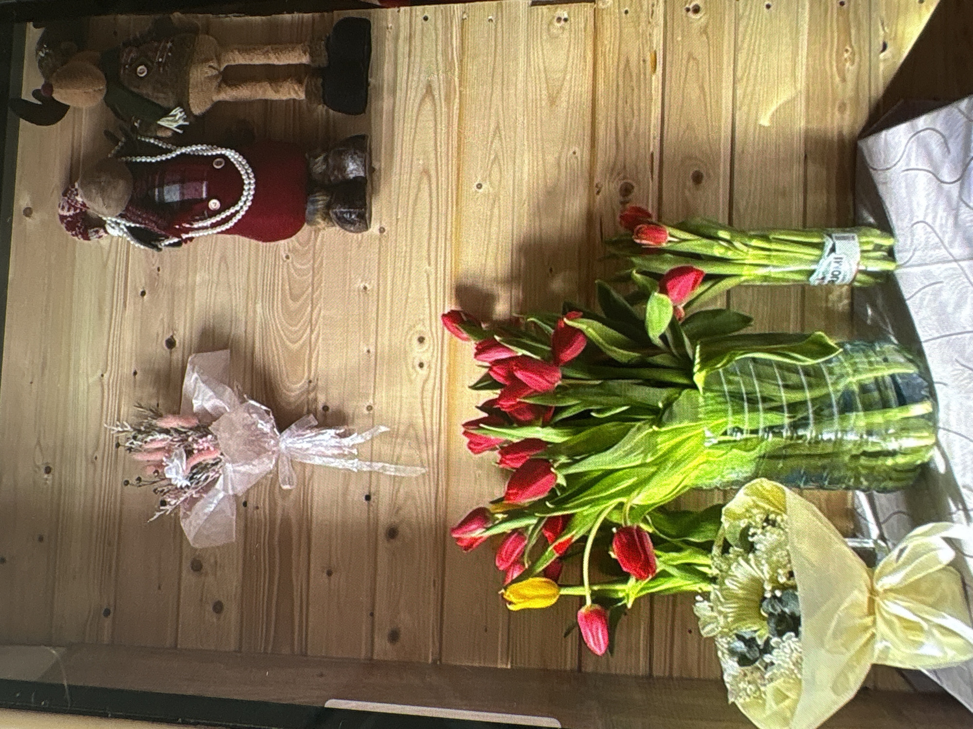 The image depicts an indoor scene with a wooden plank wall as the backdrop. In the upper left portion of the wall, a small bouquet of dried flowers, wrapped in sheer pink material and tied with a ribbon, is mounted. To the right of this, two plush stuffed moose figures are displayed. The larger moose on the left wears a red garment, a checkered scarf, and a pearl necklace, while the smaller moose on the right is dressed in a brown vest and a green scarf.
Below the wall decorations, on a surface covered with a light-colored tablecloth featuring a subtle pattern, are three bouquets of tulips. The largest bouquet, predominantly red with one yellow tulip, is arranged in a clear, textured vase on the right. To the left of this, a smaller bouquet of red tulips stands in a tall, narrow clear vase, which has a label that reads "SUOMI". In the far left foreground, a smaller bouquet of white flowers, possibly daisies and baby's breath, is wrapped in pale yellow paper and tied with a yellow ribbon.
The lighting suggests it is daytime, with sunlight casting shadows on the wall. The scene appears to be a decorative display, possibly in a home or a small shop. There are no people present in the image, and no specific identifying features of the city of Batumi, Georgia, are visible.
