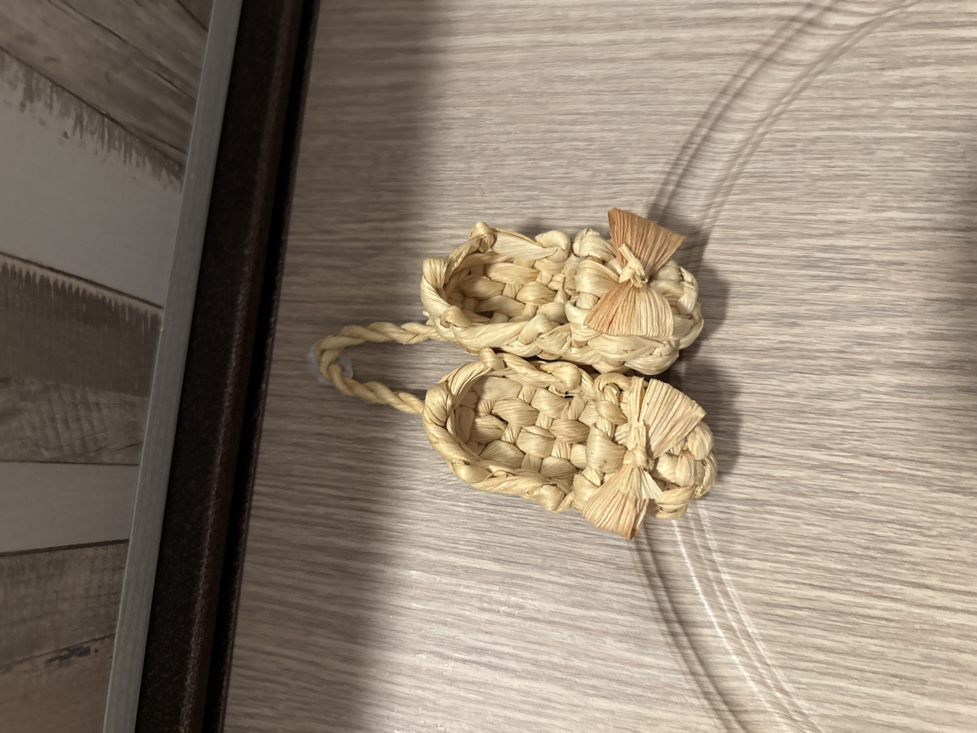 The image displays two miniature, decorative sandals, hand-woven from light-toned natural fibers, likely straw. Each sandal features a tightly braided construction with a small, delicate bow made of the same material affixed to the toe. The sandals are connected at their heels by a braided loop, indicating they are hung from an unseen fixture.
They are positioned against a vertical surface exhibiting a light-colored, linear wood-grain pattern, characteristic of a laminated door or wall panel. Above this panel, a dark brown, horizontal textured strip, consistent with a door frame or trim, spans the upper width of the image. Above the dark strip, a lighter grey horizontal element is visible, followed by another panel with a distinct wood-grain pattern, suggesting an indoor setting with architectural detailing.
Subtle shadows are cast by the woven sandals onto the background panel, indicating a primary light source positioned above and slightly to the left of the frame. A faint, curved shadow is also discernible on the lower right portion of the main wood-grain panel.
No discernible details within the image, such as specific architectural features, unique objects, or text, allow for the identification of the city of Yekaterinburg, Russia, or any other specific geographical location. The scene depicts generic indoor decorative items and architectural elements.