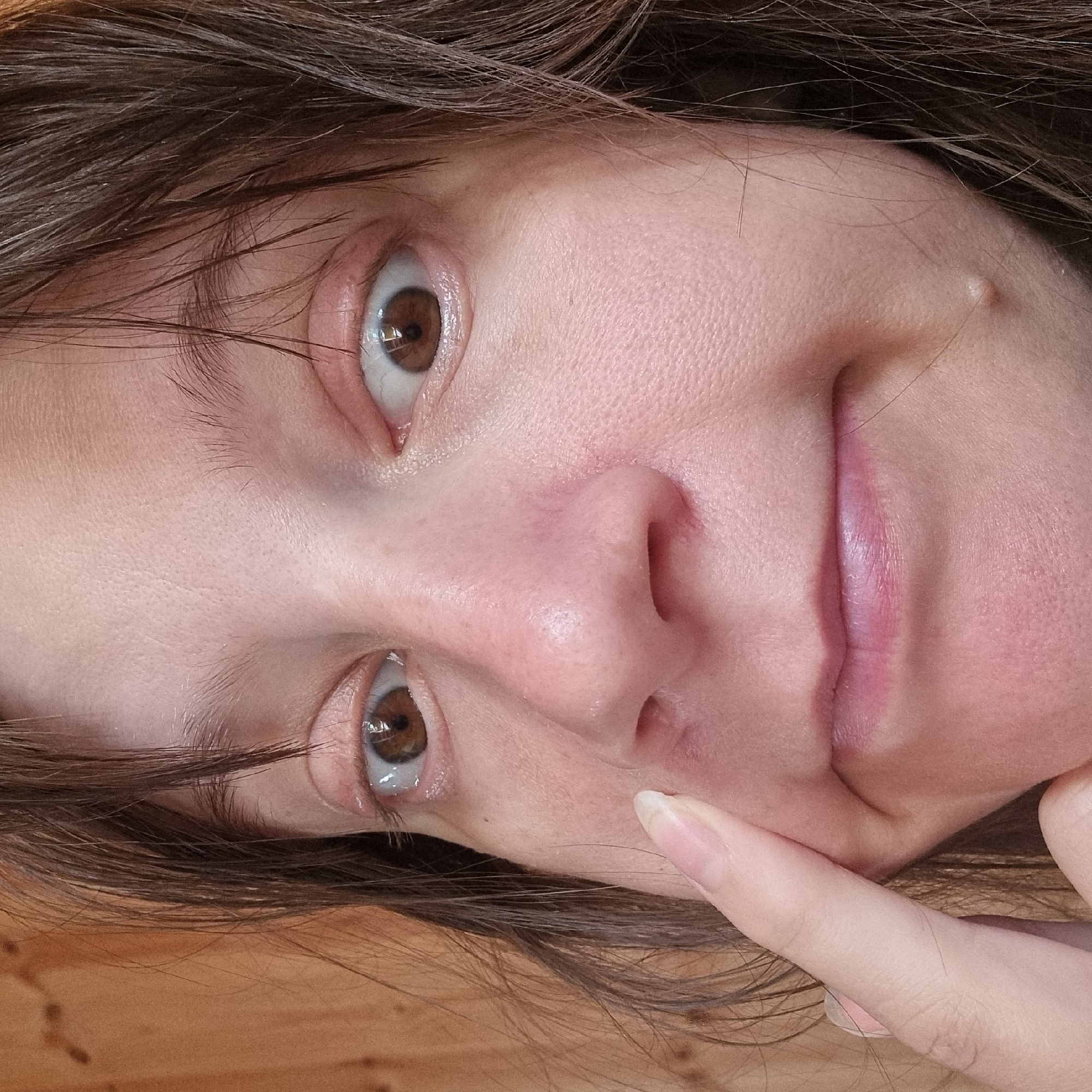 The image shows a close-up of a person's face, focusing on their eyes, nose, and mouth. The person has brown eyes and light skin. Their hair is dark brown and appears to be somewhat messy. A finger is visible in the left side of the frame, touching the cheek near the nose. There is a small, raised bump on the person's right cheek. The background is out of focus but appears to be a wooden surface. There is no information available about the location in Dunnae-myeon, South Korea.