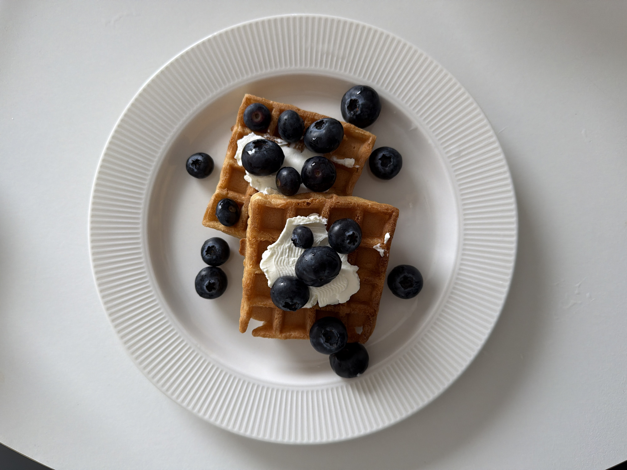 The image displays a top-down view of two golden-brown square waffles positioned on a white, circular plate with a fluted rim. Each waffle is topped with a distinct dollop of white cream, appearing to be whipped cream, and multiple dark blue blueberries. Additional blueberries are scattered on the plate's surface around the waffles. The plate rests on a flat, plain white surface, likely a table. The scene is evenly lit, with no discernible shadows indicating a direct light source. No individuals or contextual environmental details beyond the immediate tabletop are visible. This scene is located in Tel-Aviv, Israel.