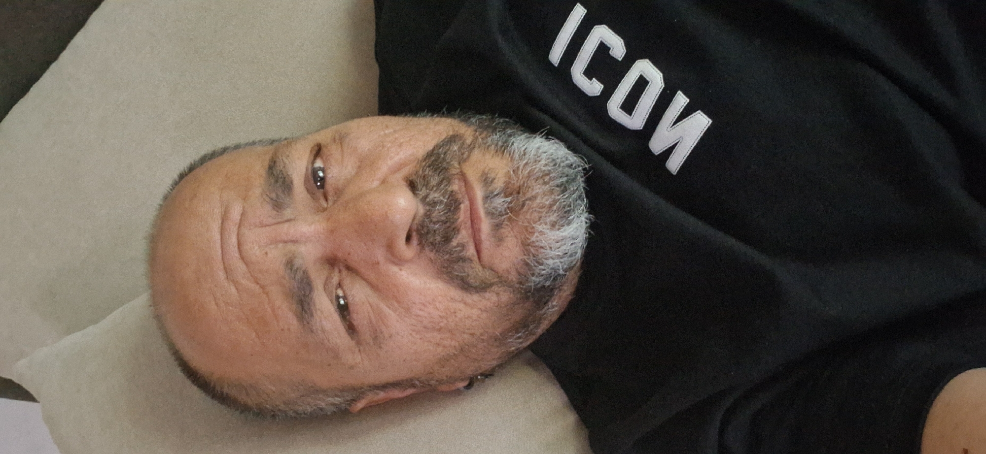 The image is a portrait of a man, possibly located in Simeonovgrad, Bulgaria, lying down indoors. His head is turned towards the camera, and he is resting on a light beige or grey cushion. He appears to be a middle-aged or older man with a fair to medium complexion, characterized by a salt-and-pepper beard and short, dark hair with visible grey streaks. His eyes are open, and he maintains a neutral or slightly thoughtful expression.
He is wearing a black shirt or sweatshirt, prominently featuring the word "ICON" in large, white, outlined letters across the chest area. The fabric of the shirt shows some natural creases. The lighting is soft and ambient, suggesting an indoor setting, likely indicating a moment of rest or relaxation.