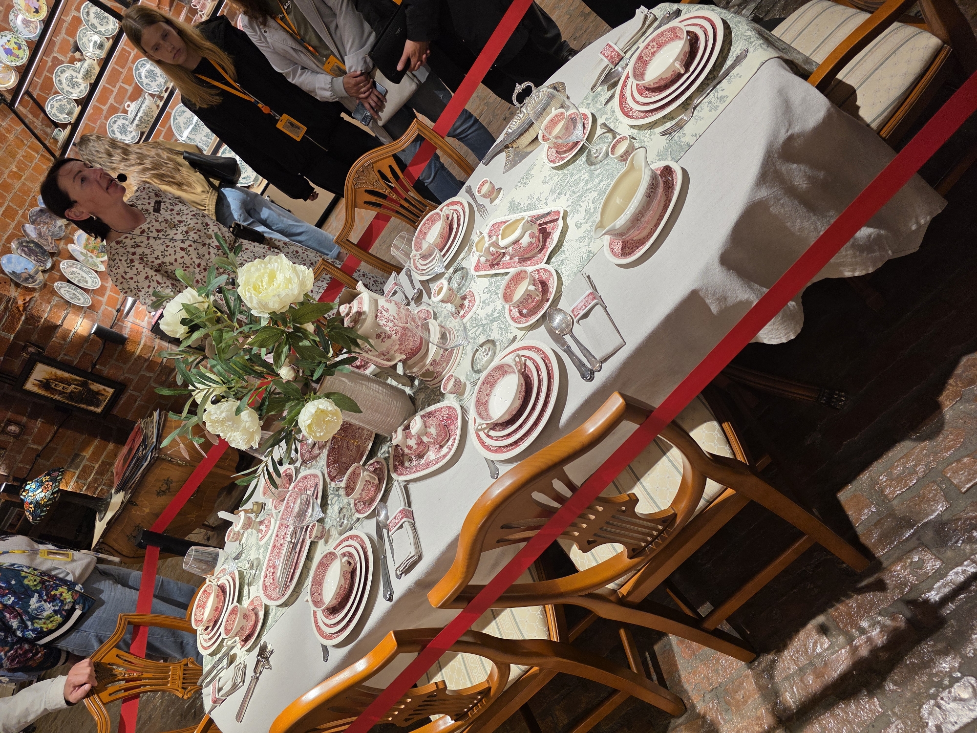 The image depicts a museum setting, likely an exhibition of decorative arts, featuring a meticulously set dining table with a collection of pink and white patterned porcelain china. The table is covered with a white tablecloth and a runner with a floral pattern. On the table, there are numerous cups, saucers, plates, a teapot, cream pitcher, gravy boat, and other serving dishes, all part of the same china set. The arrangement suggests a formal tea or dining service.
In the background, several people are visible, observing the display. Some are wearing lanyards with badges, indicating they might be museum staff or visitors on a guided tour. The environment appears to be indoors, with exposed brick walls and shelves displaying more decorative plates. A Tiffany-style lamp is visible on a wooden chest in the background. The overall impression is one of a historical or cultural exhibit.
While the image itself does not explicitly state its location, if this exhibition is indeed in Kaliningrad, Russia, a potential context could be a museum like the Kaliningrad Regional Museum of Art or a historical estate showcasing period furnishings and decorative arts. The style of the china and the overall presentation are consistent with exhibits found in such institutions.