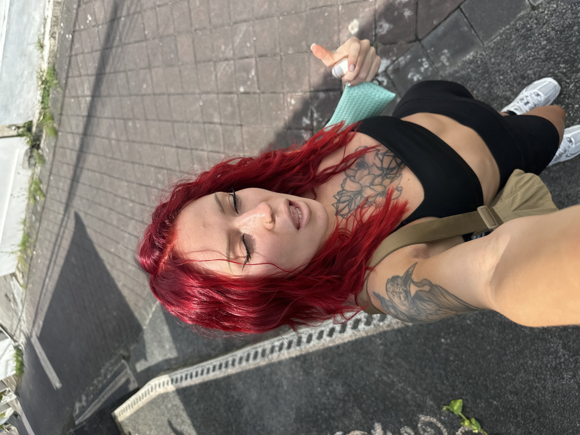 A woman with red hair and tattoos takes a selfie in North Kuta, Indonesia. She's wearing a black top and black shorts, with white sneakers. She is holding a small, white object in her left hand, with a turquoise object underneath. She's standing on a grey stone walkway. A khaki-colored bag is slung over her shoulder. Shadows suggest it's a sunny day.